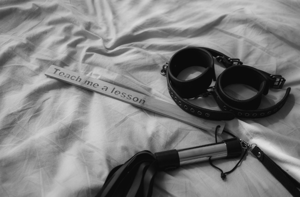 Een houten plank met de tekst 'Teach me a lesson', een paar zwarte handboeien en een flogger