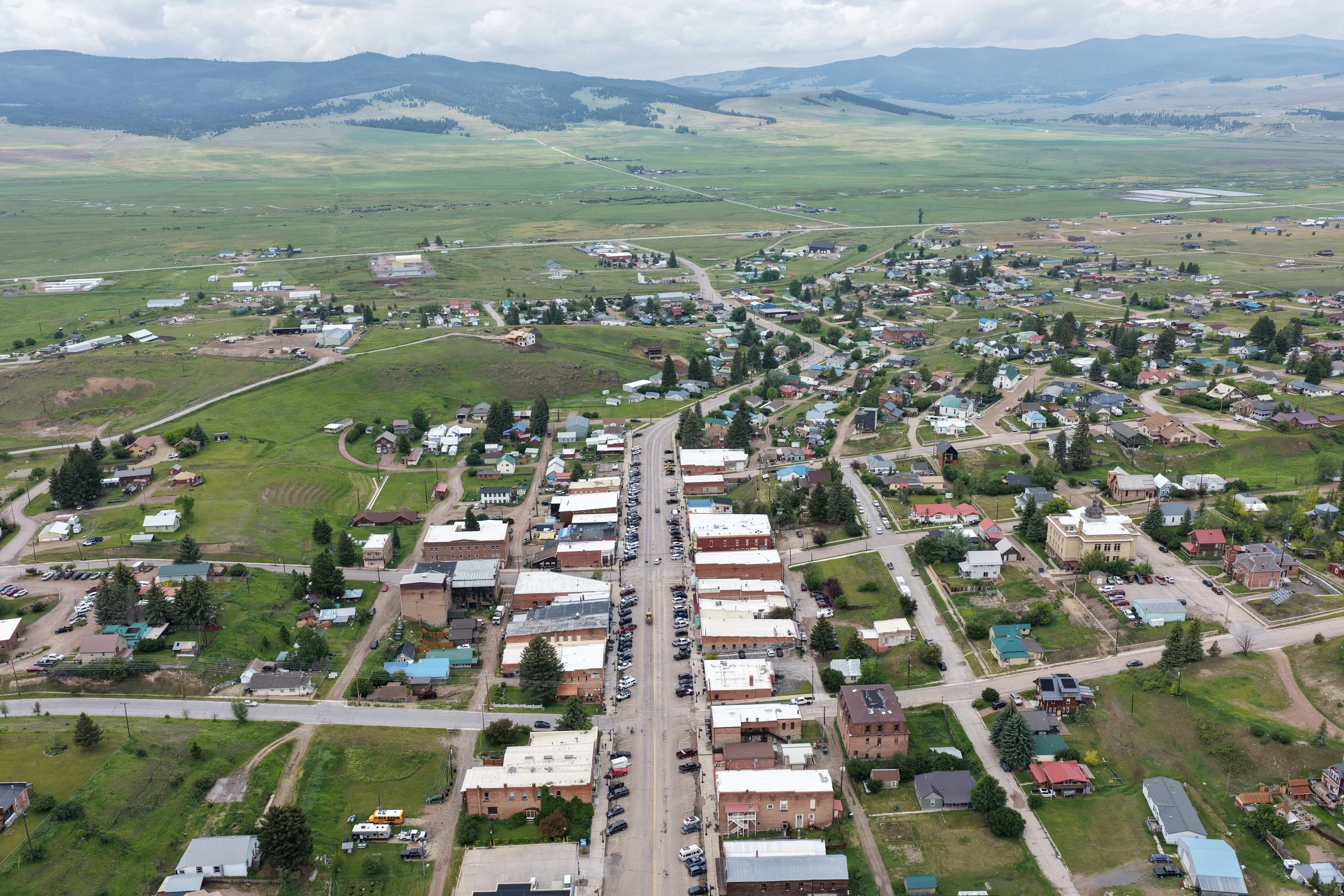 Martin aerial drone view of philipsburg montana with a mix of residential and commercial buildings surrounded by green fields and hills in the background under a cloudy sky.
