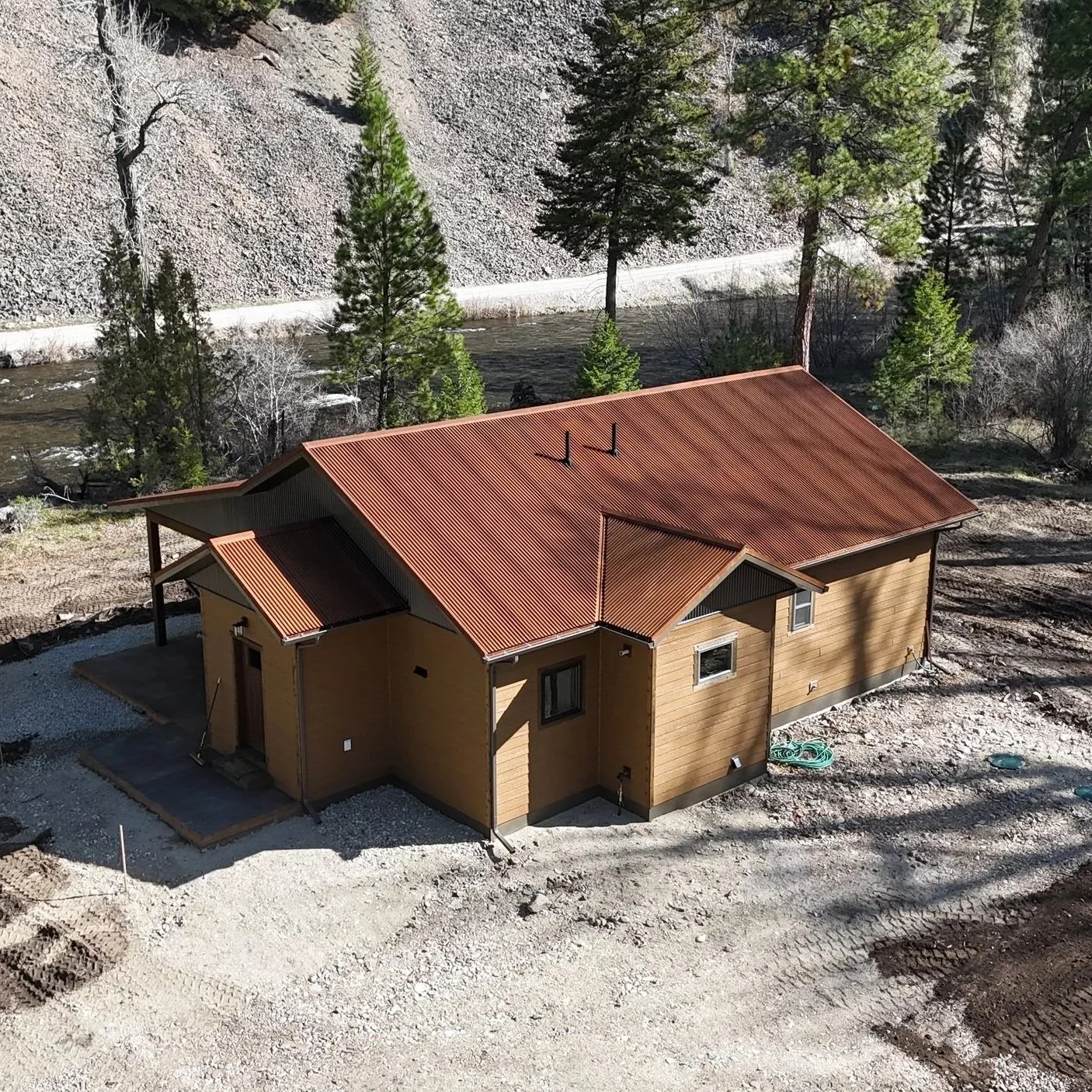 Martin aerial drone A house with a metal roof under construction, situated in a wooded area near a river, with trees and a rocky hillside in the background.