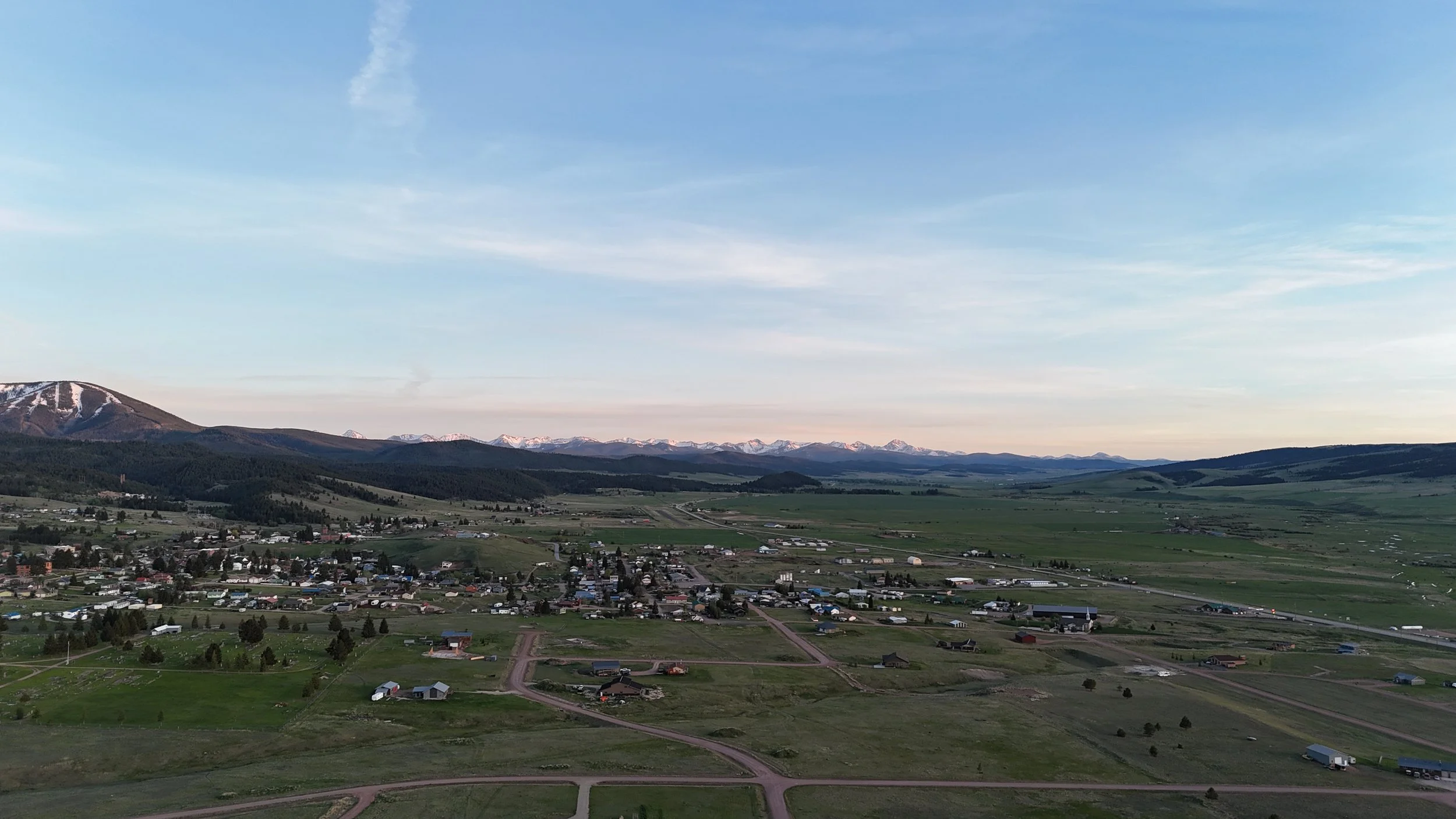 Martin aerial drone A scenic landscape with a view of small town buildings, green farmland, rolling hills, and snow-capped mountains under a clear blue sky.