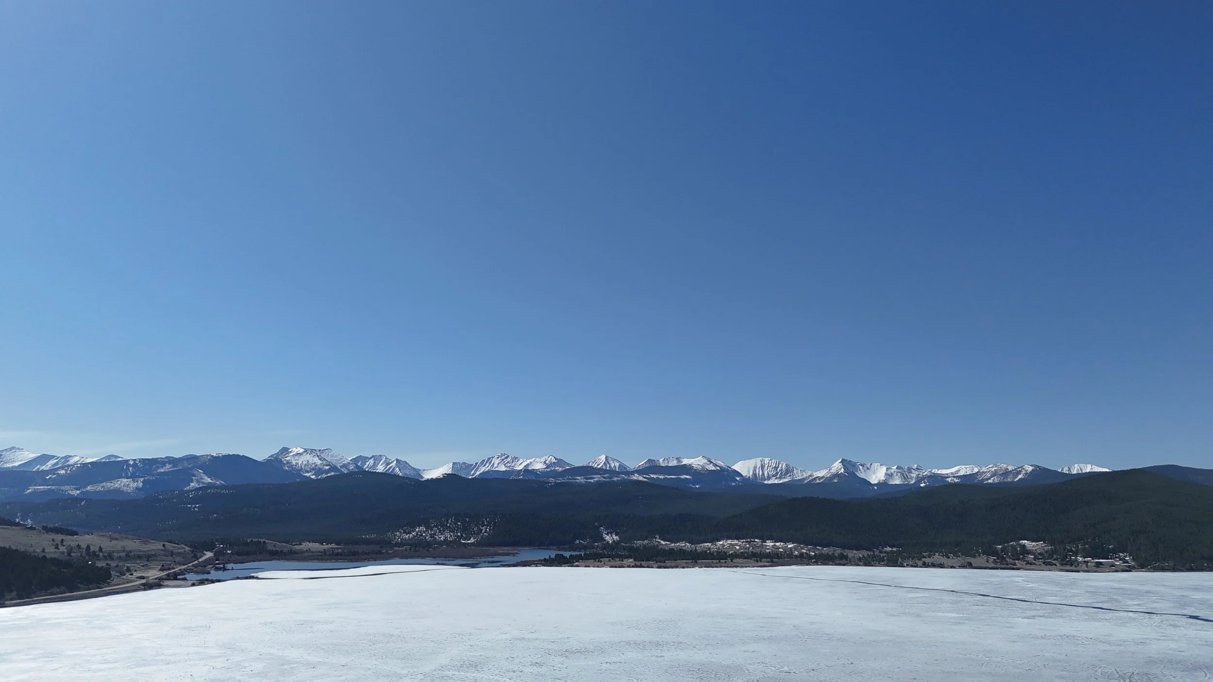 Martin aerial drone Snow-covered landscape with a mountain range in the background and a large open frozen body of water in the foreground under a clear blue sky.