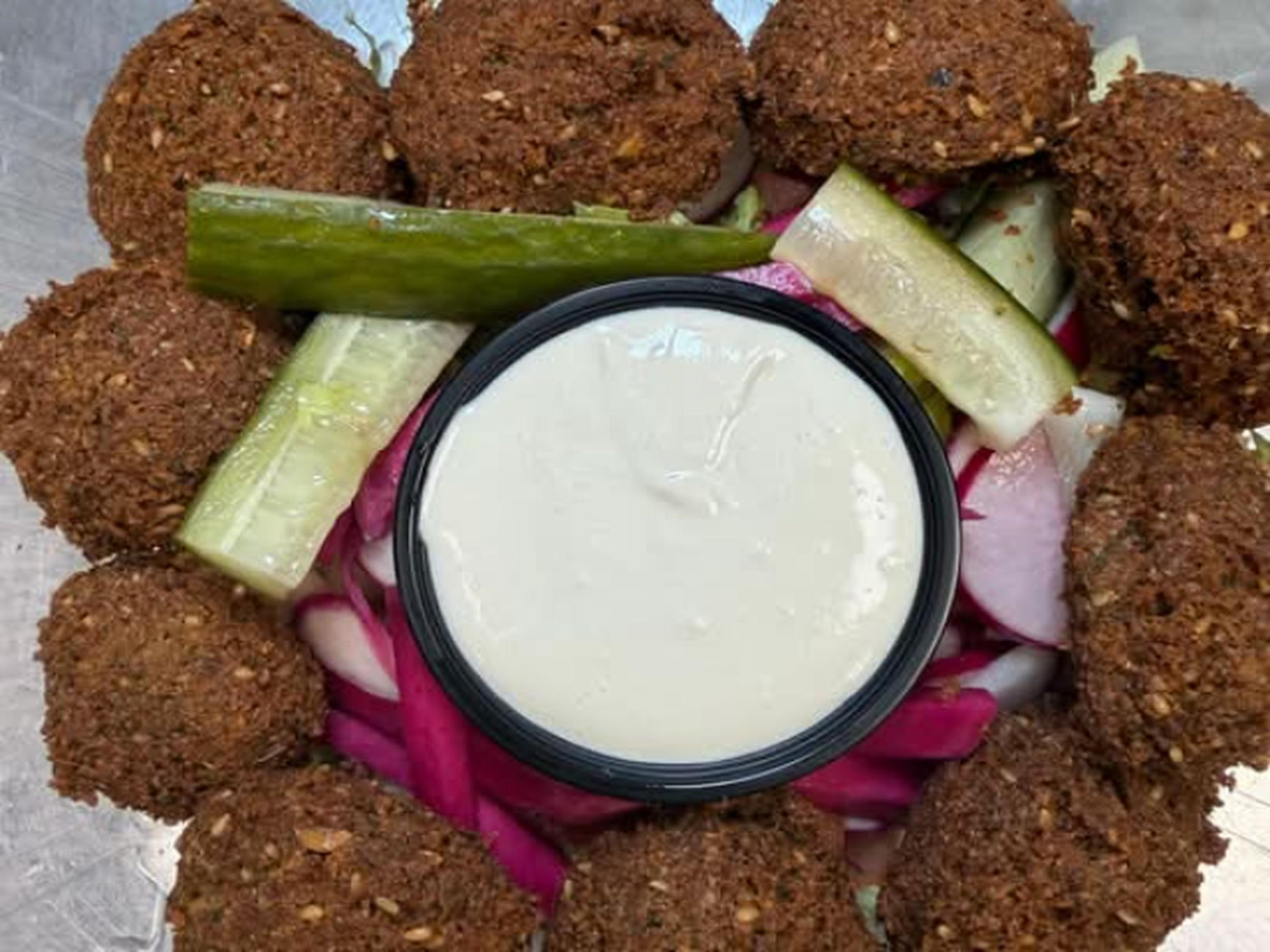 A person wearing a pink glove holding a small bowl of creamy white sauce or dressing, surrounded by large falafel balls and sliced cucumbers.