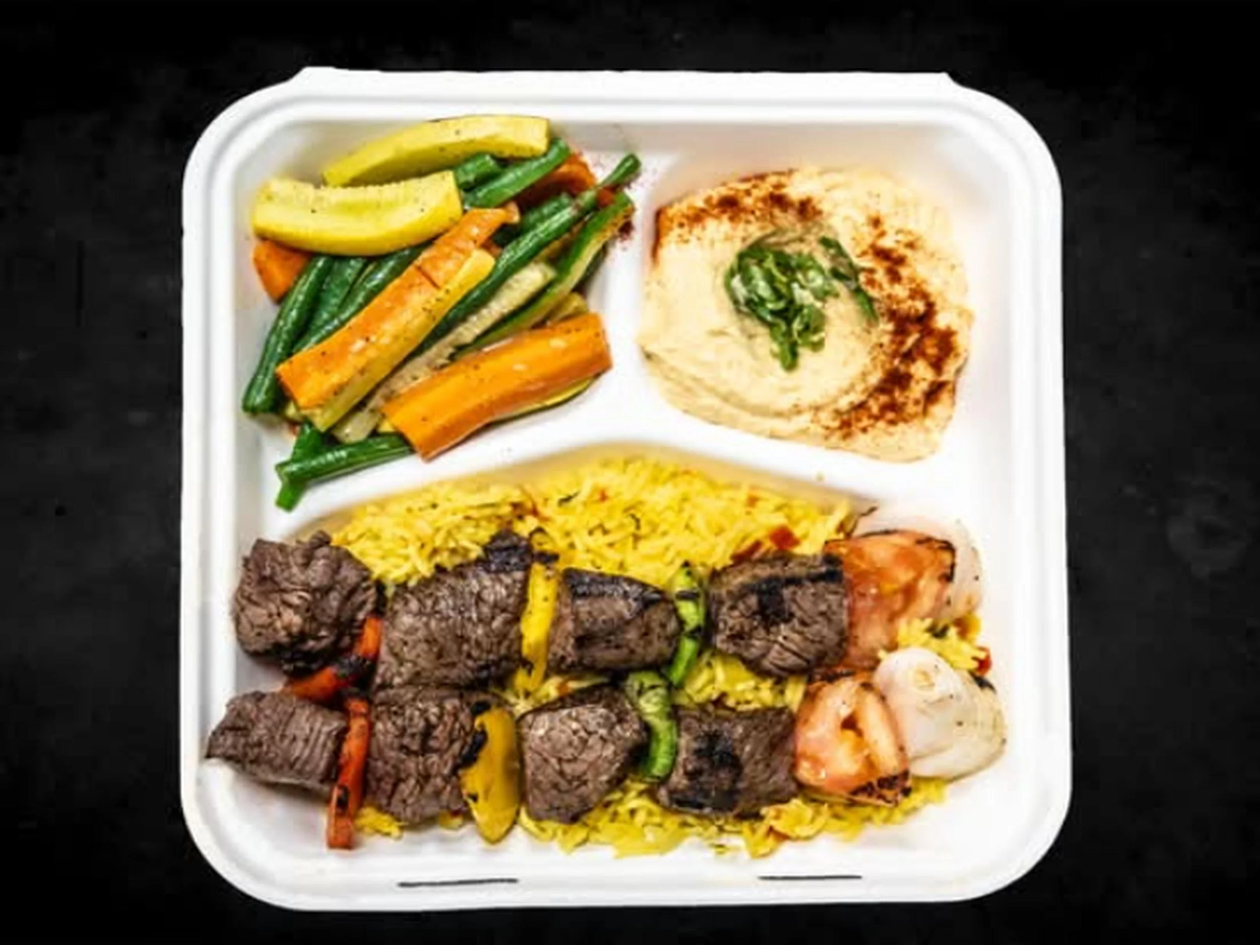 A white compartmentalized foam takeout container holding grilled meat, yellow rice, and vegetables including green beans, carrots, and zucchini.