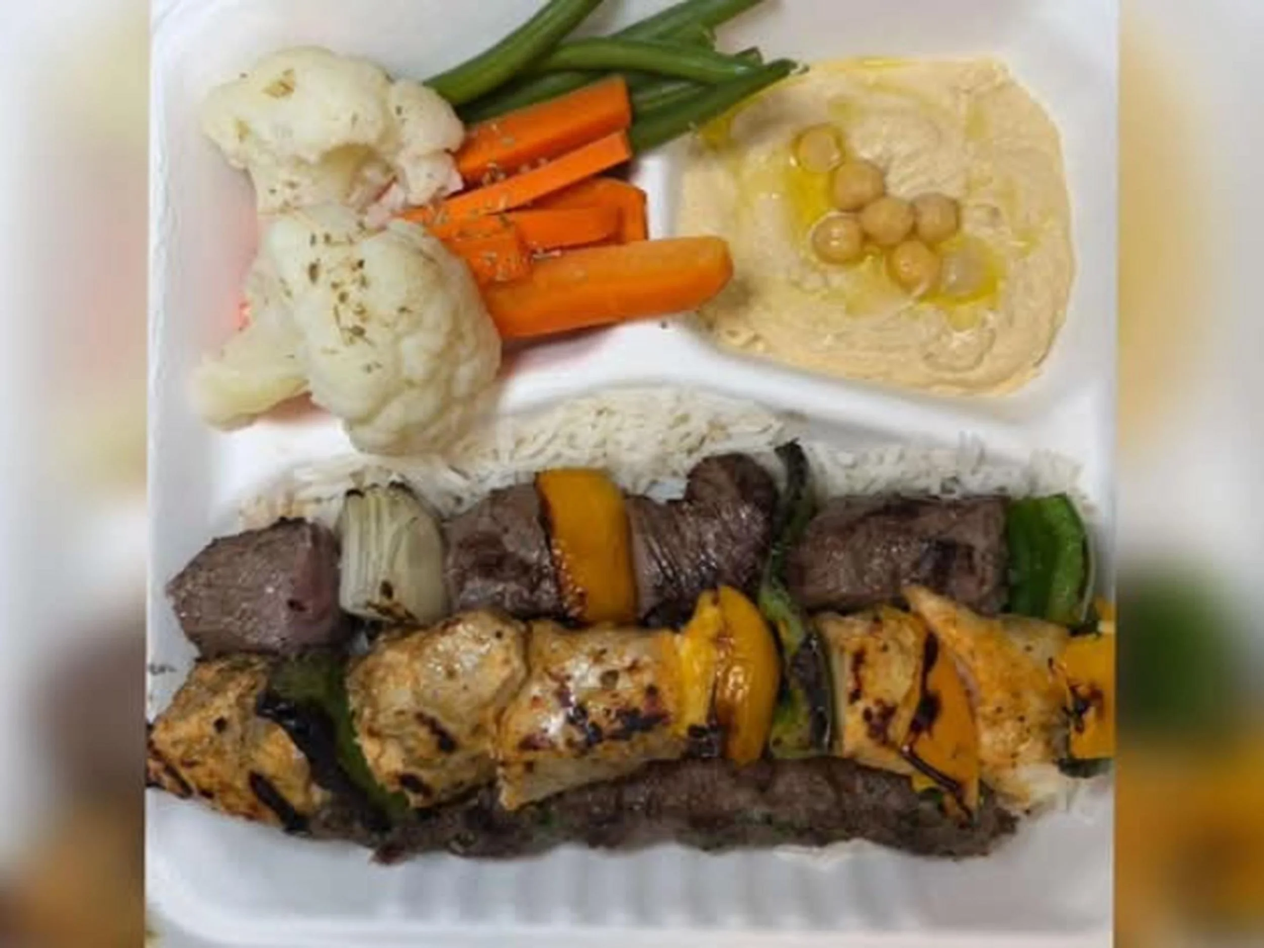 A takeout box filled with grilled vegetables and beef, including cauliflower, carrots, green beans, and chunks of steak.
