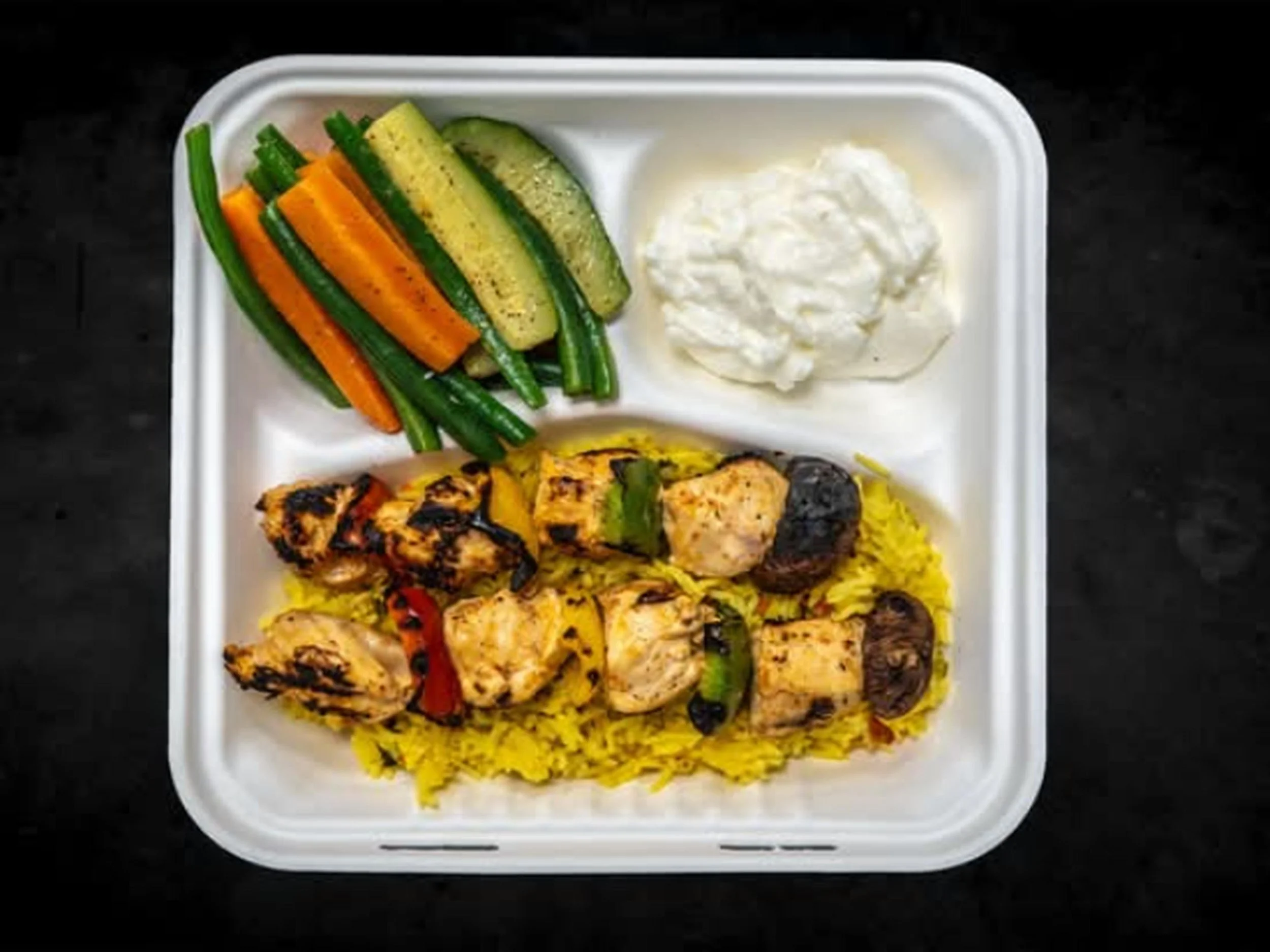 A white takeout container with grilled chicken skewers, yellow rice, grilled vegetables, fresh cucumber slices, carrot sticks, green beans, and a side of creamy white dressing or sauce.