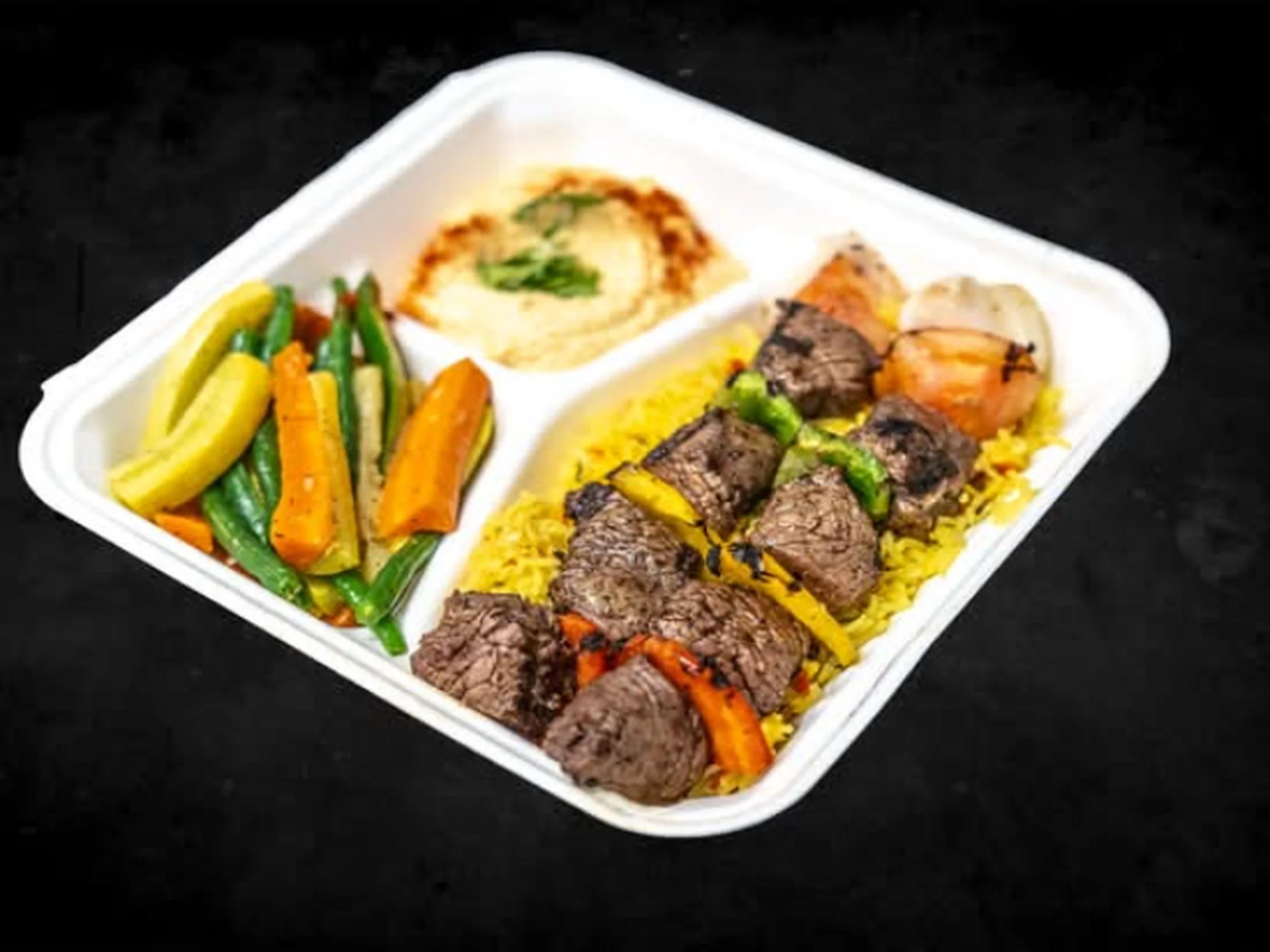 A divided white foam plate containing cooked vegetables, a scoop of mashed potatoes with green herbs, grilled beef pieces, and sliced cooked carrots.