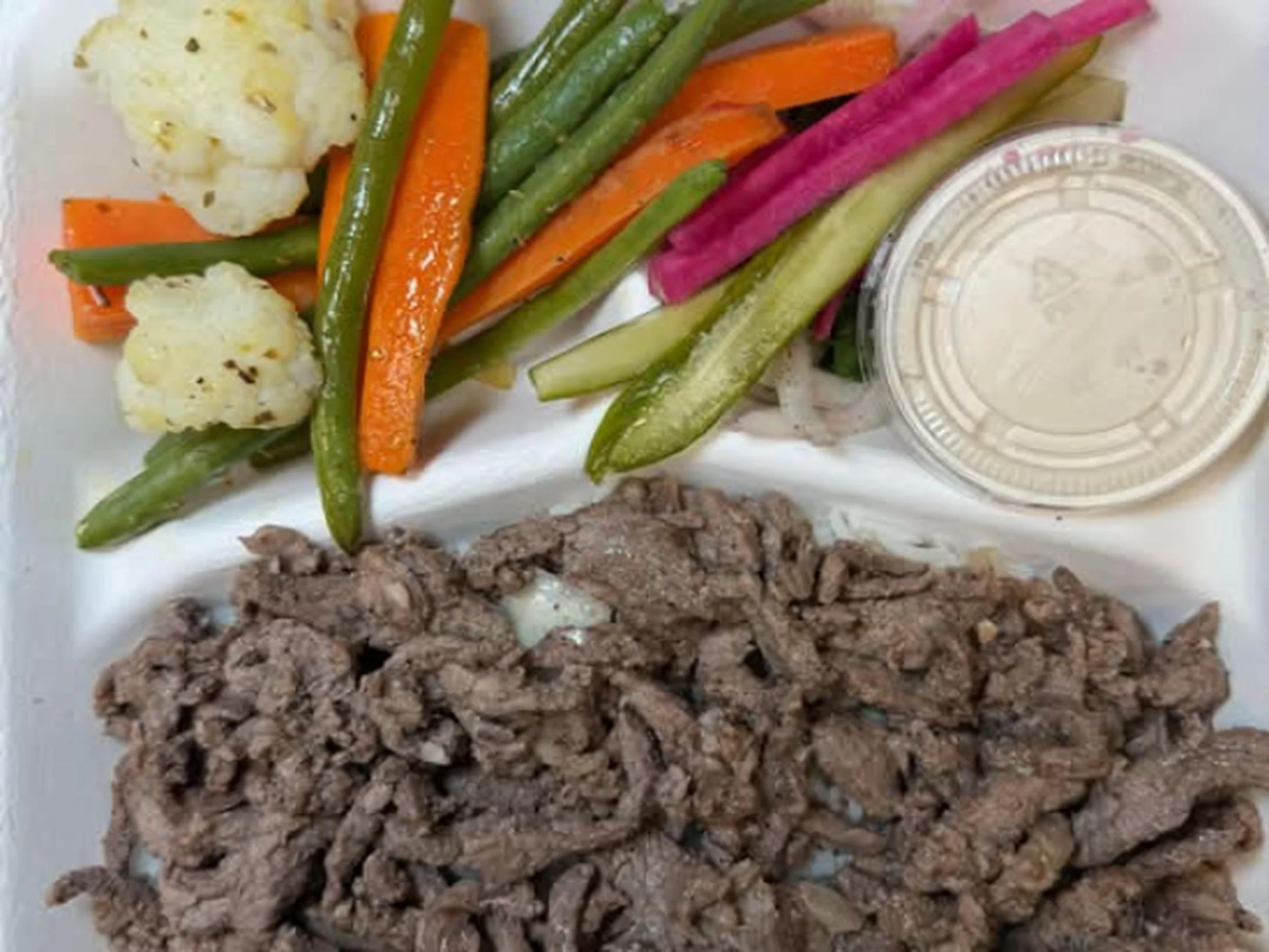 A plate of cooked vegetables including cauliflower, green beans, carrots, pink and green sweet potato fries, a small container of dipping sauce, and ground beef.