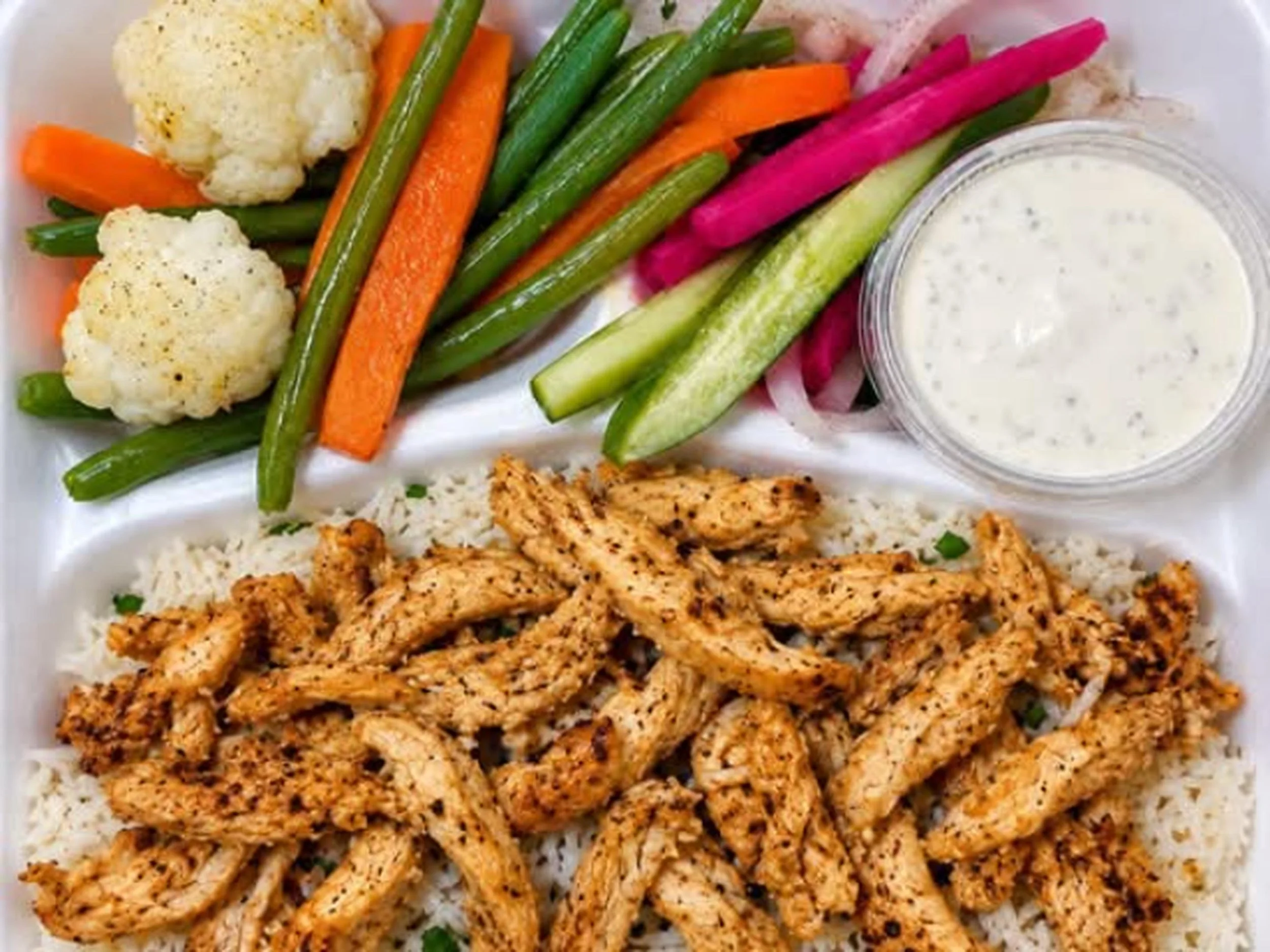 Grilled chicken strips served over rice with assorted fresh vegetables including cauliflower, carrots, green beans, and cucumber, accompanied by a side of creamy dipping sauce.