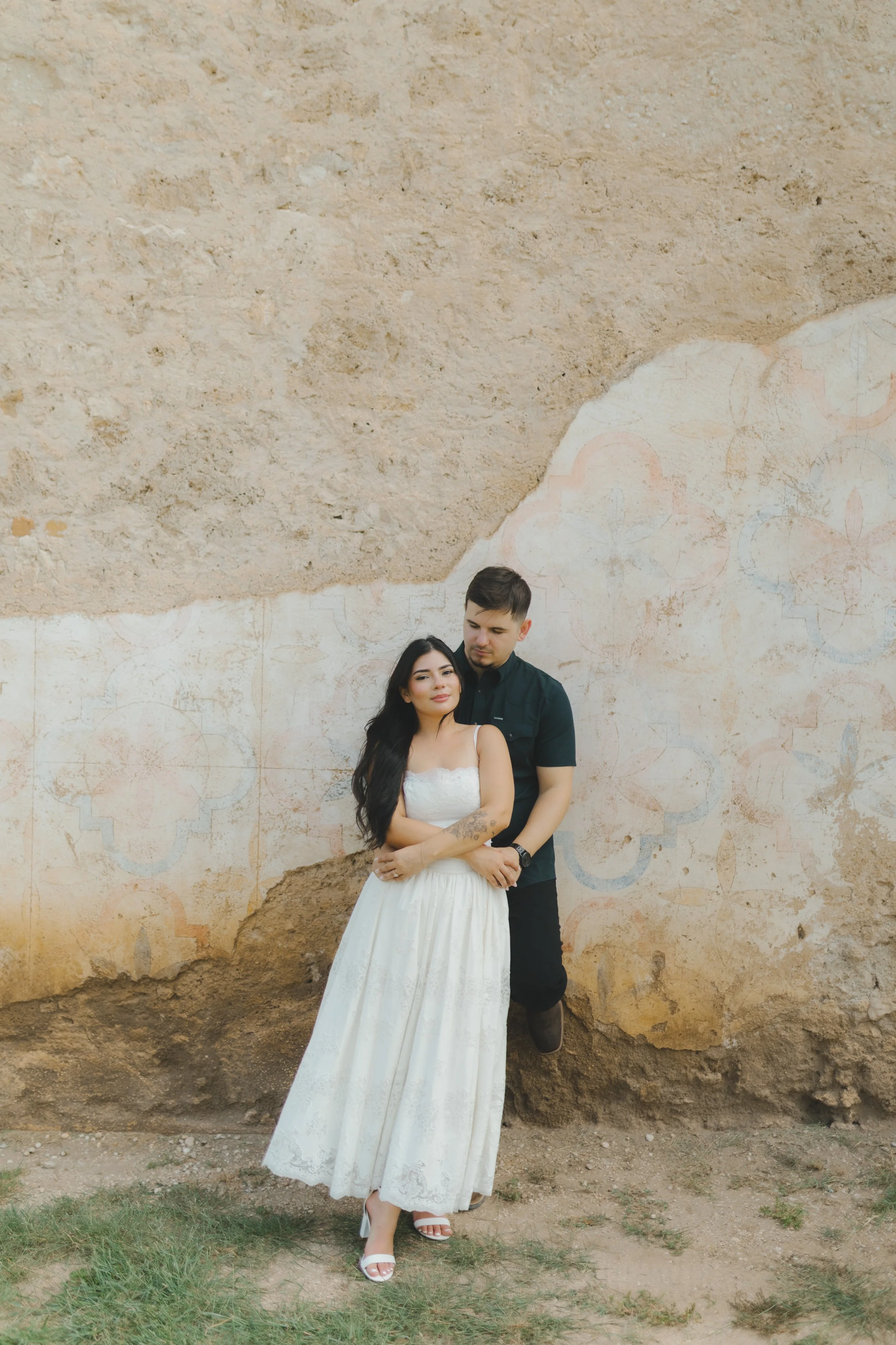 A young woman in a white wedding dress and a young man in dark clothing standing against an aged wall with painted designs. The woman has long dark hair, and the man is looking down at her, embracing her from behind.