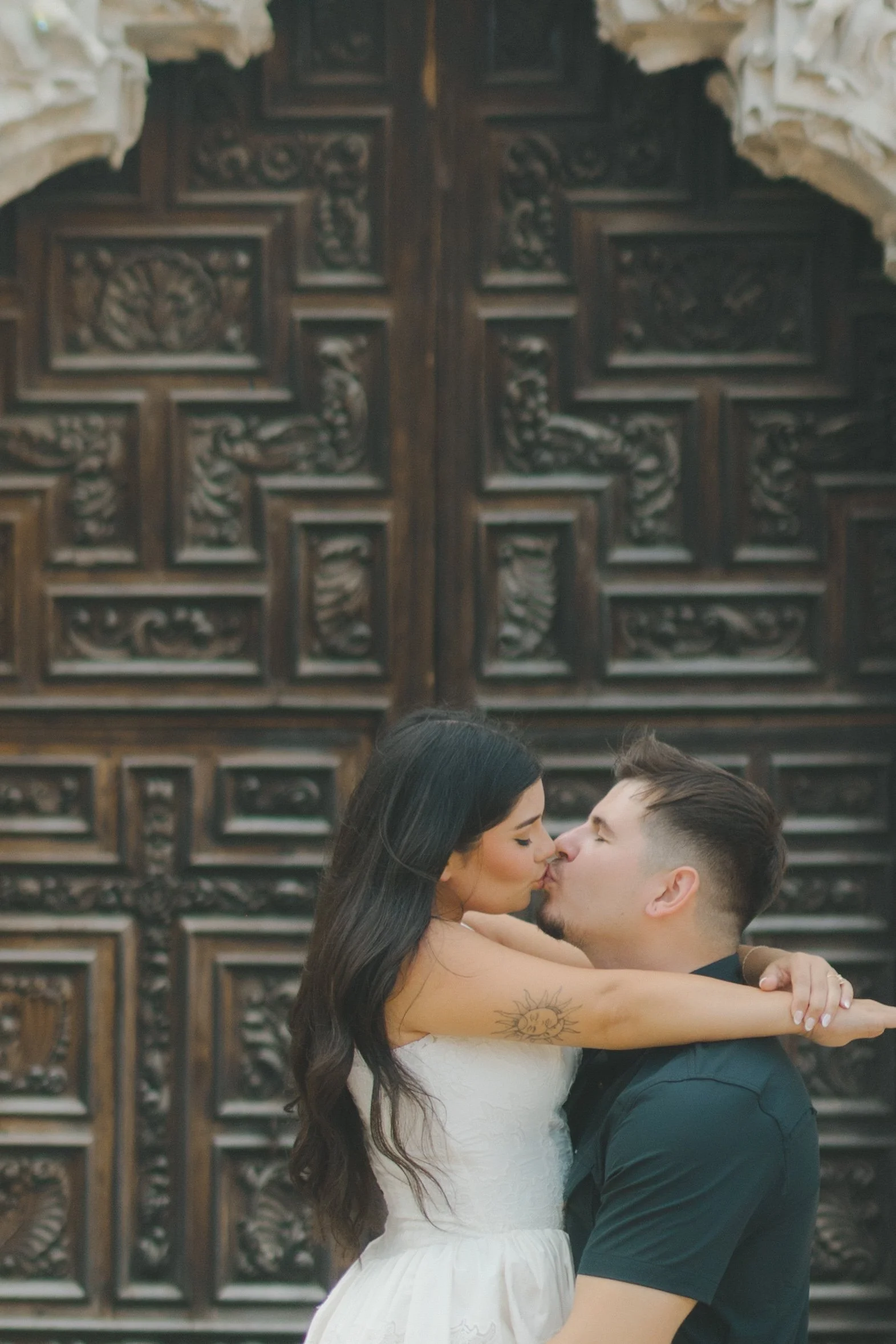 A couple sharing a kiss in front of a decorative wooden panel, the woman with long dark hair and a tattoo on her arm, and the man with short hair and a goatee, both embracing each other.
