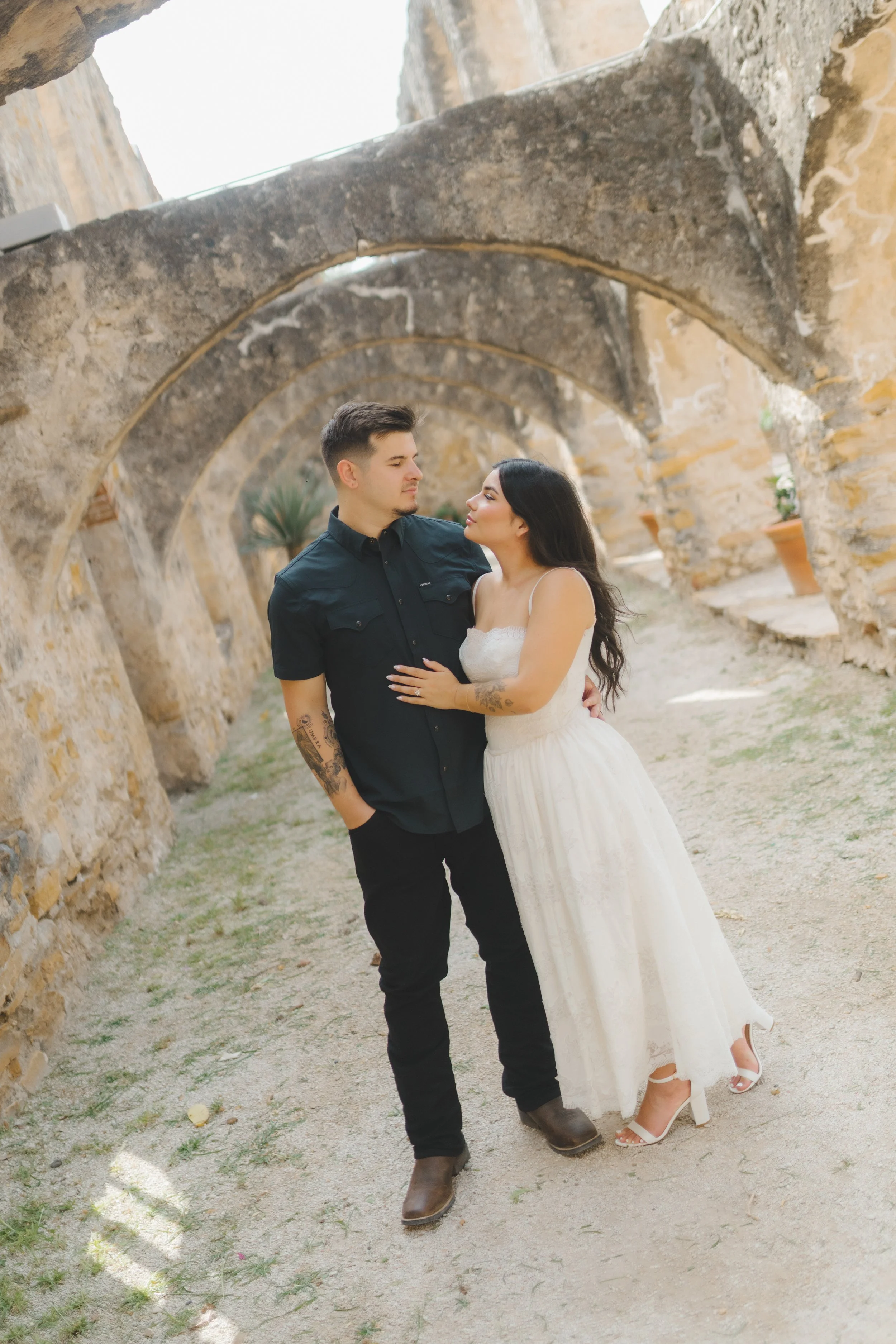 A man and woman standing close together in a stone archway, with the woman in a white dress and the man in black shirt and pants, looking at each other