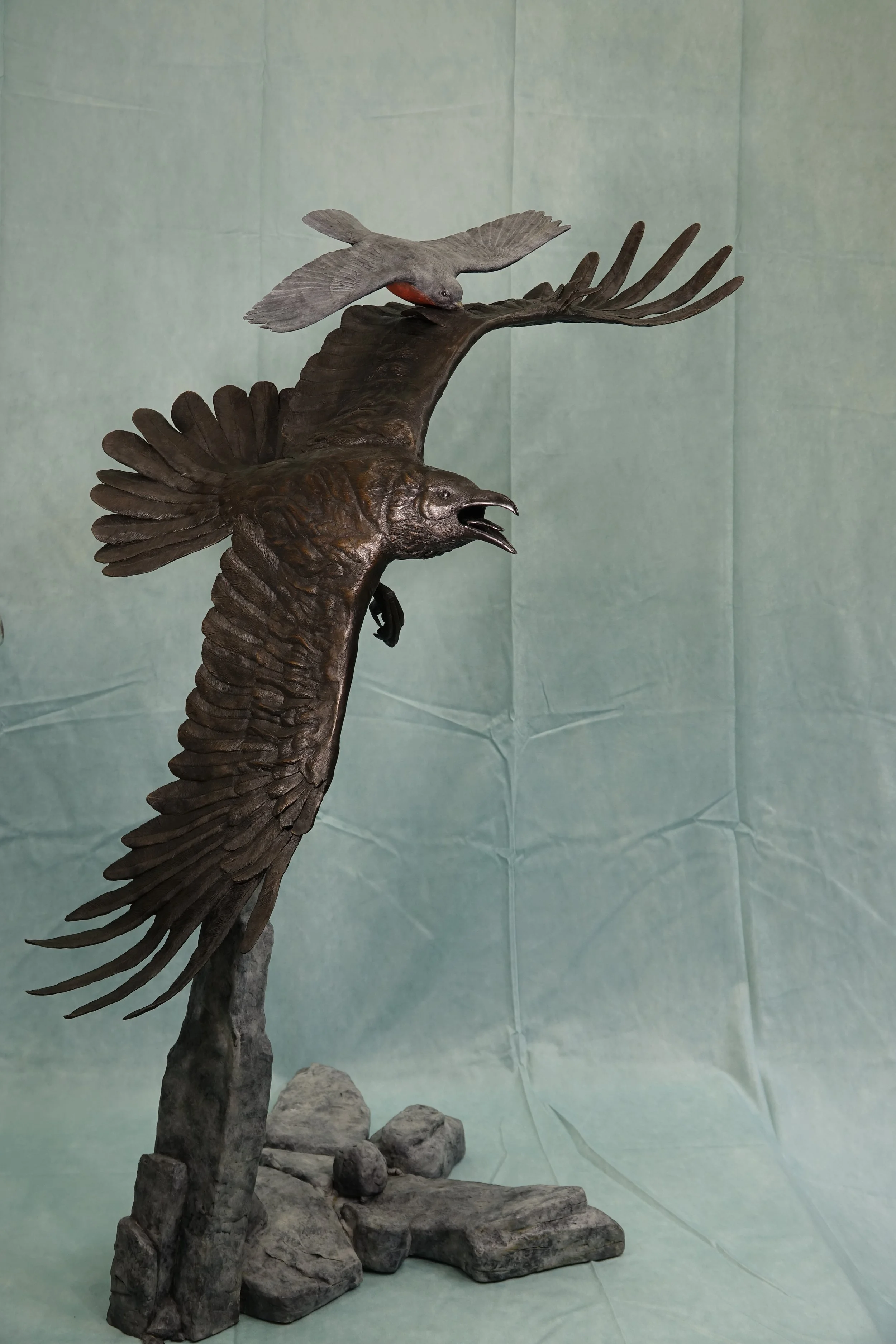 Statue of an eagle with its beak open, perched on a rock, with a bird perched on a branch above.