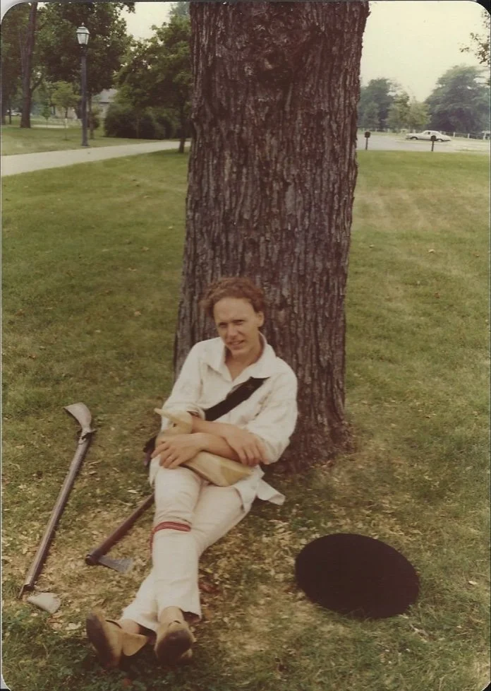 A woman sitting on the grass leaning against a large tree trunk, holding a book. A black hat is on the ground nearby, and there are crutches next to her. In the background, there is a park with trees, a sidewalk, some lamps, and a road with a car.