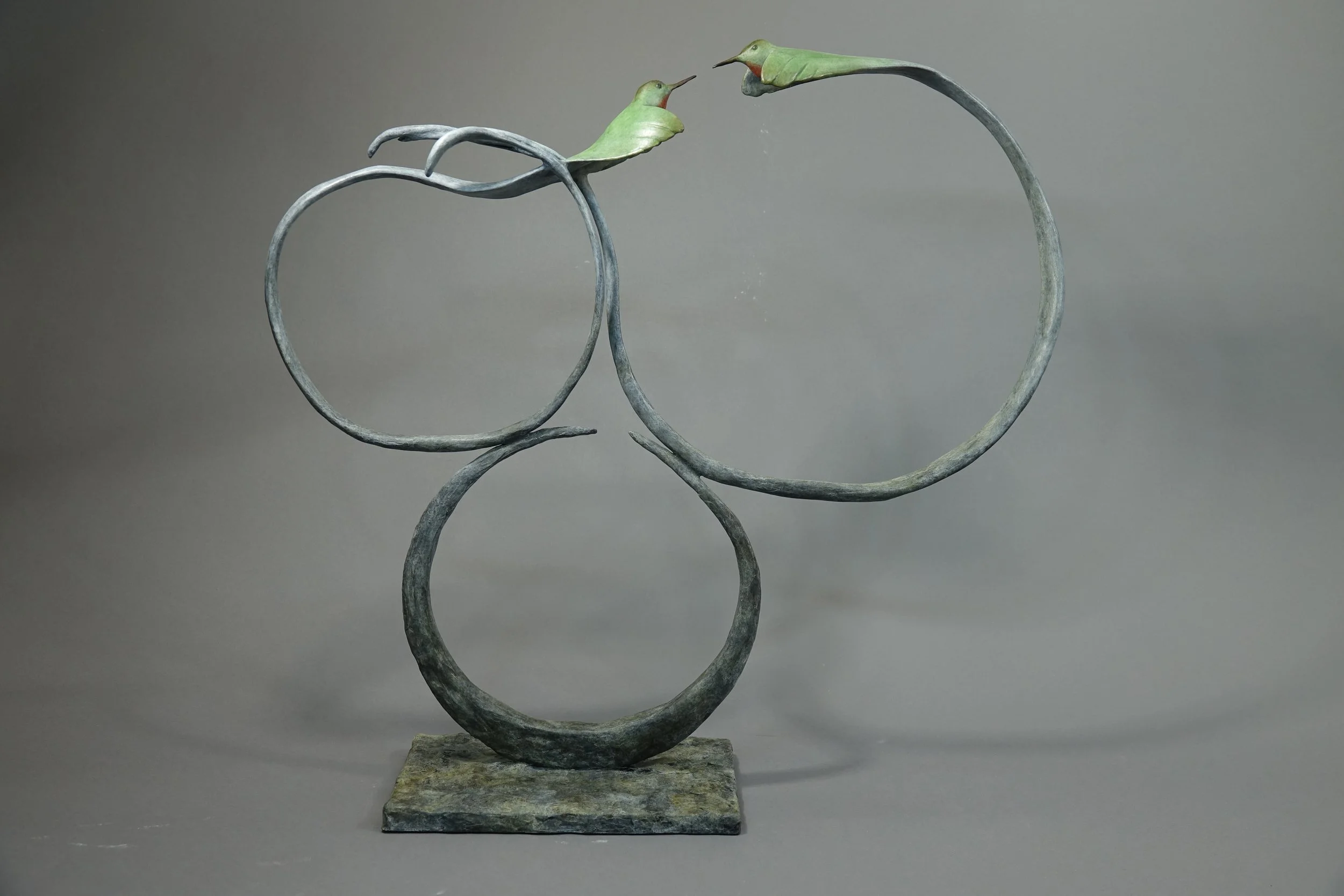A sculpture of two hummingbirds with elongated, curved stems and green leaves, facing each other, mounted on a stone base.