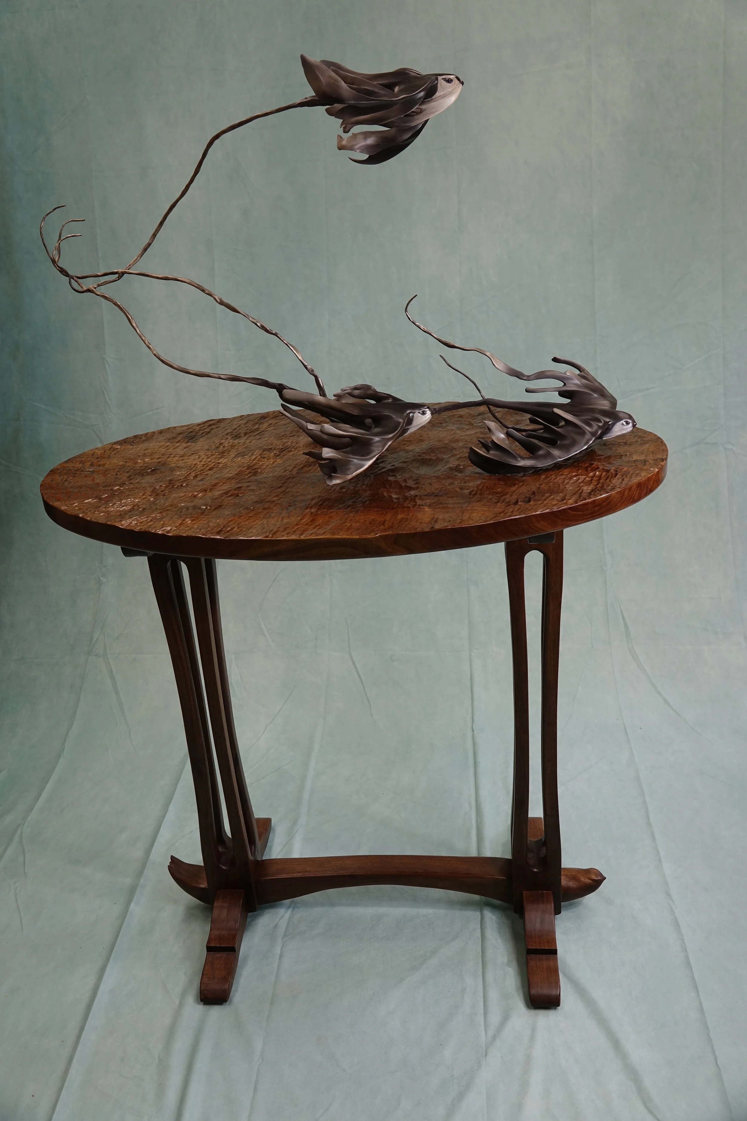 A wooden table with a unique sculpture on top, featuring dark, twisted metal and plant-like elements.