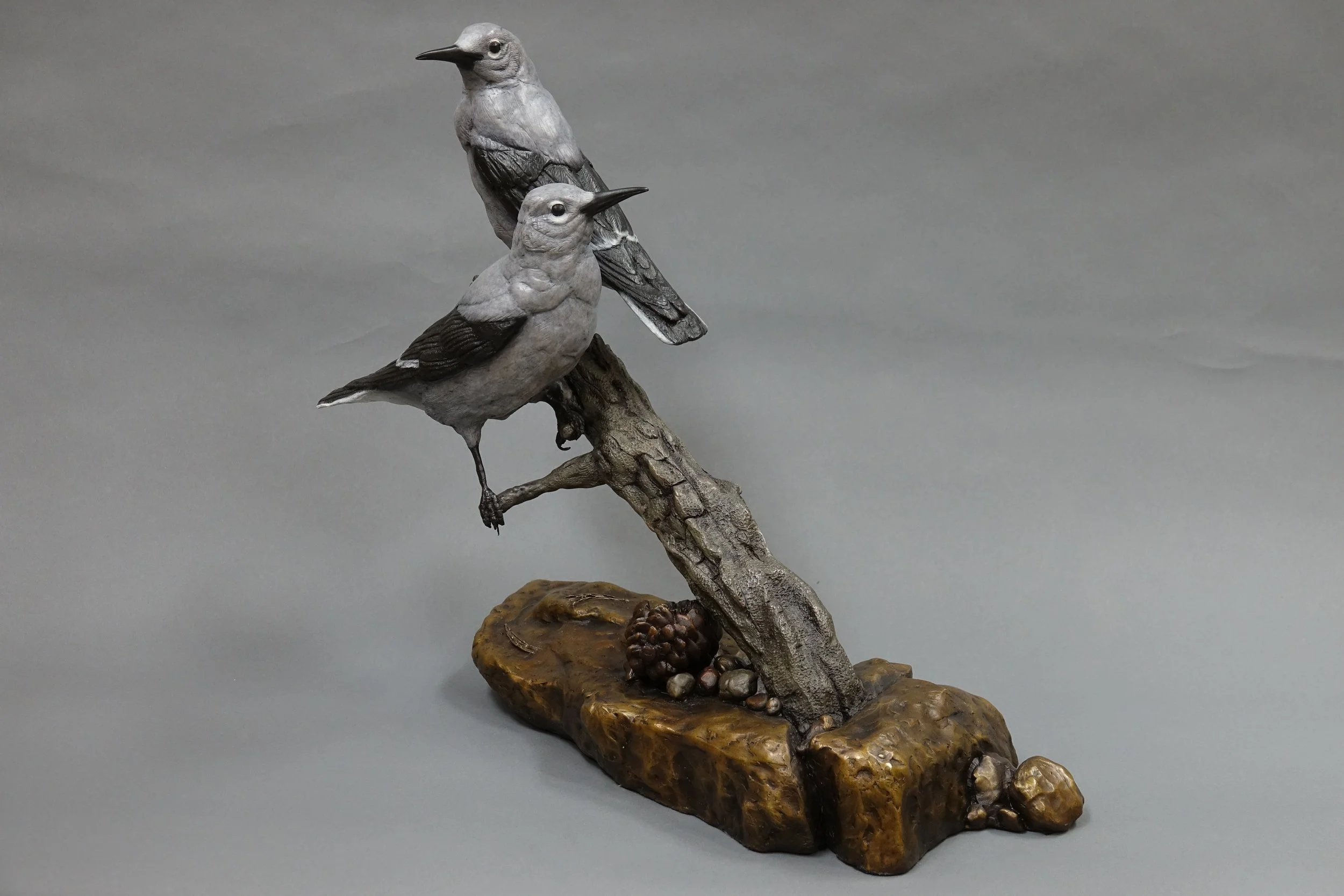 A sculpture of two birds perched on a branch, with details resembling real birds, set on a textured base with pebbles against a neutral background.