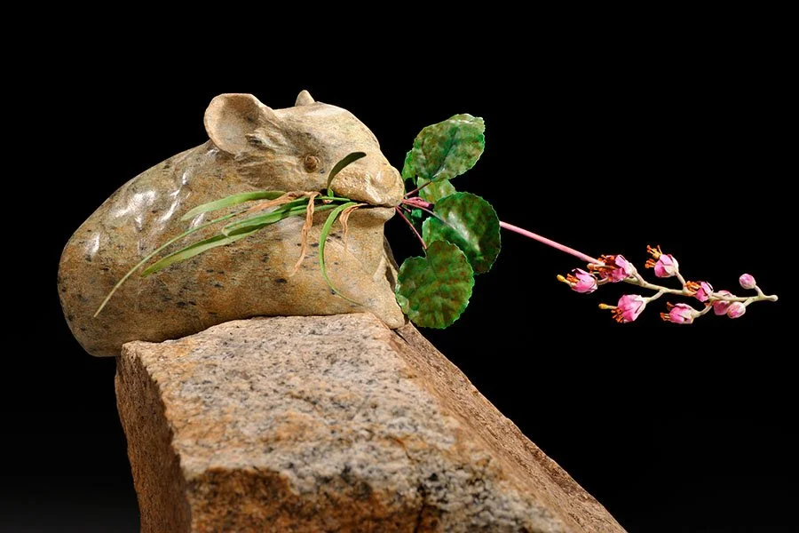 A small sculpture of a mouse made from a stone, holding a plant with pink flowers, placed on a rock against a black background.