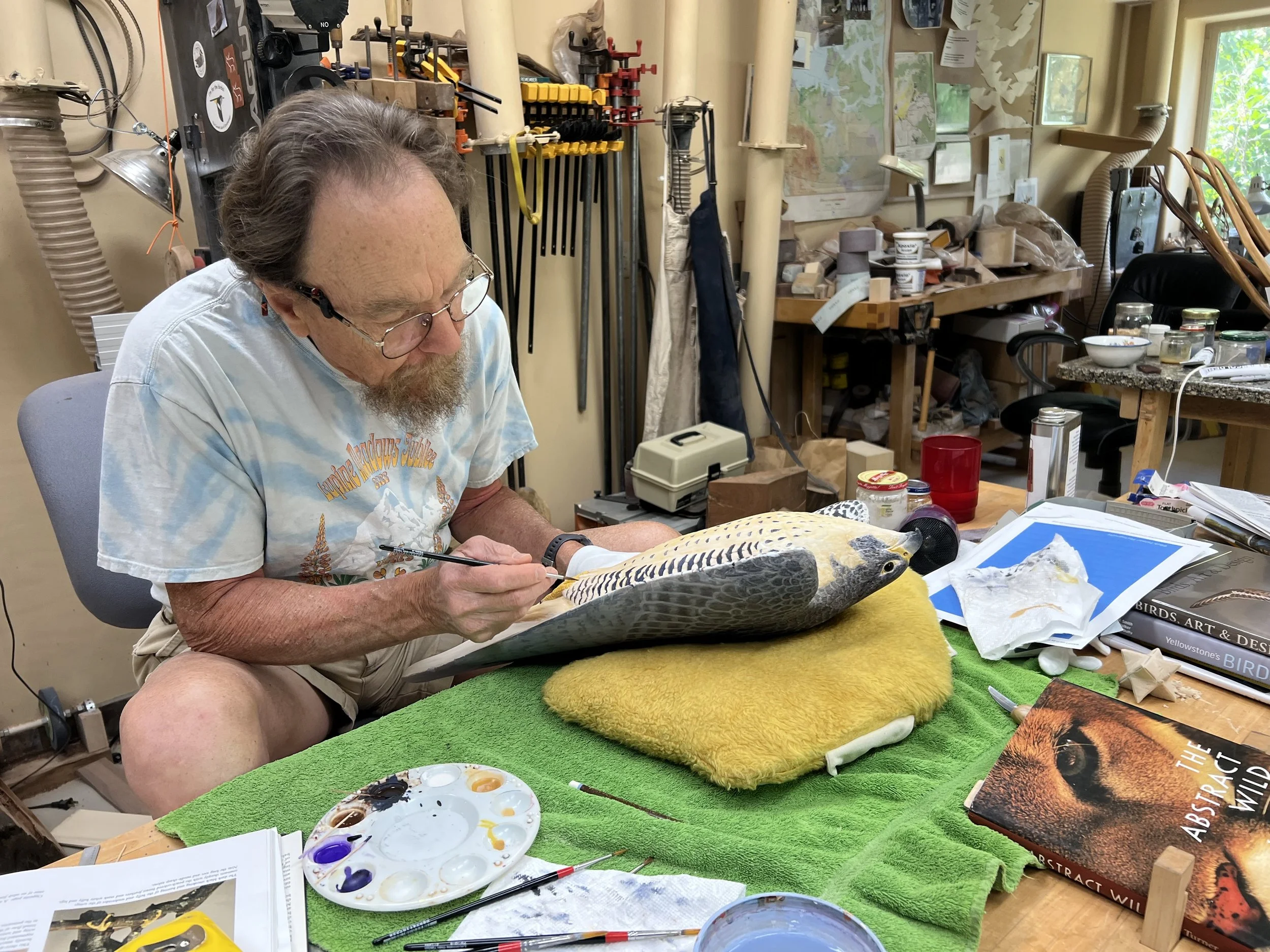 A man with glasses and a beard working on a bird specimen using a brush in a workshop filled with tools, books, and supplies.