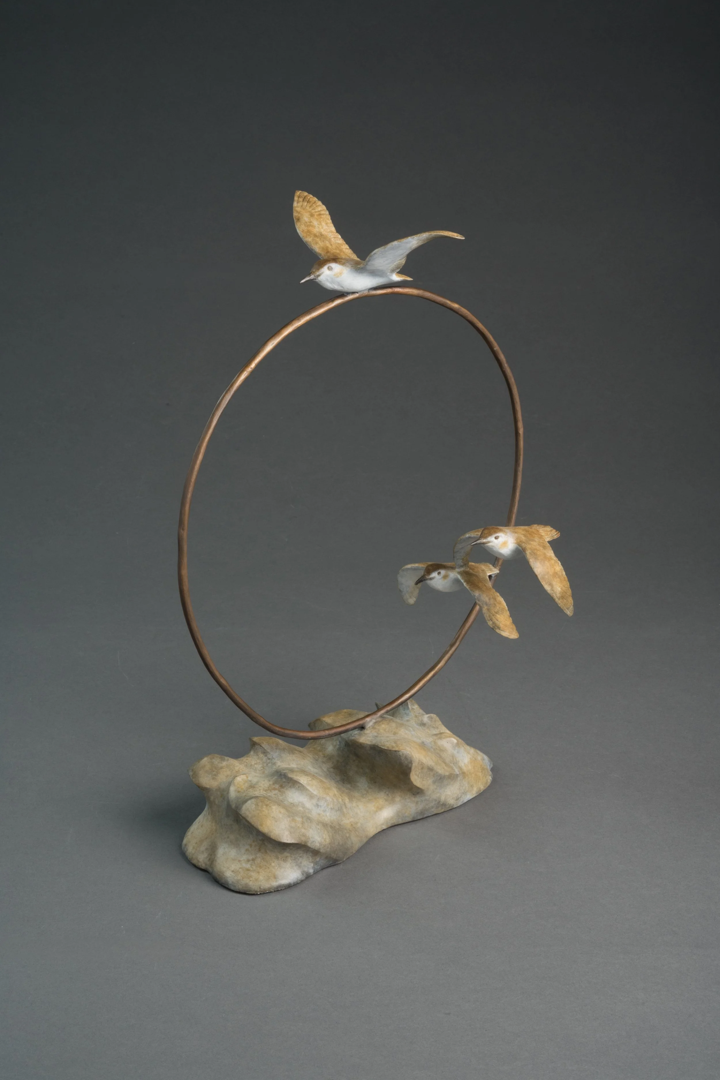 A sculpture of three birds perched on a curved metal branch, set on a large rock base, against a dark background.