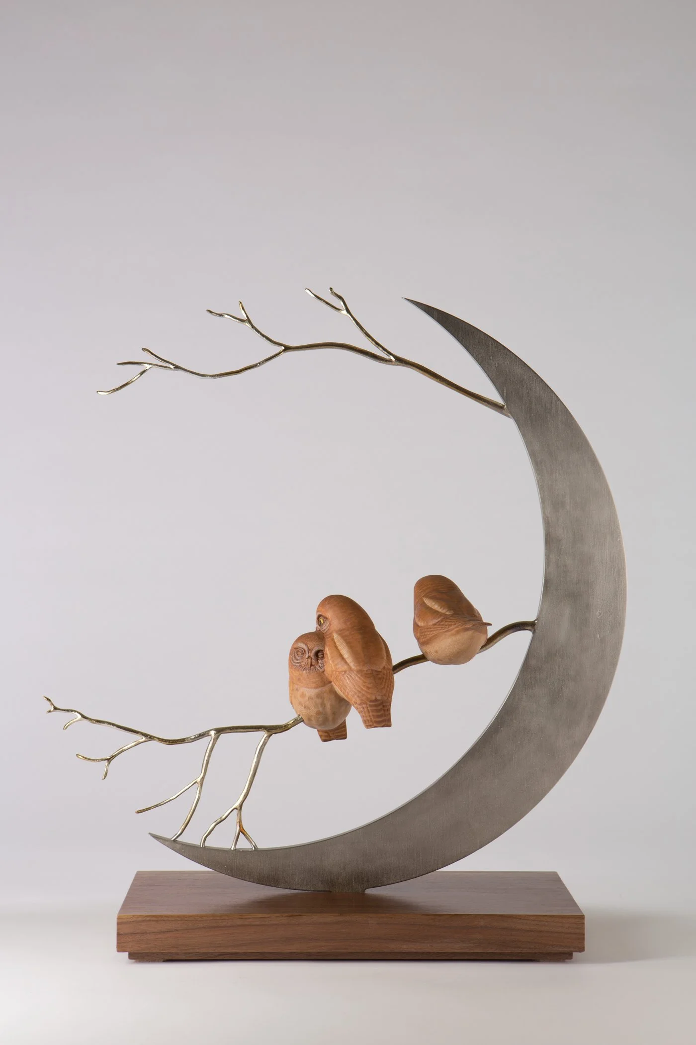 A sculpture featuring a crescent moon shape with metallic branches extending from it, holding three shells and a small plant, mounted on a wooden base.