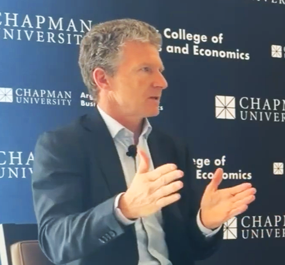 A man with gray curly hair in a suit gesturing with his hands during a presentation at Chapman University, College of Business and Economics.