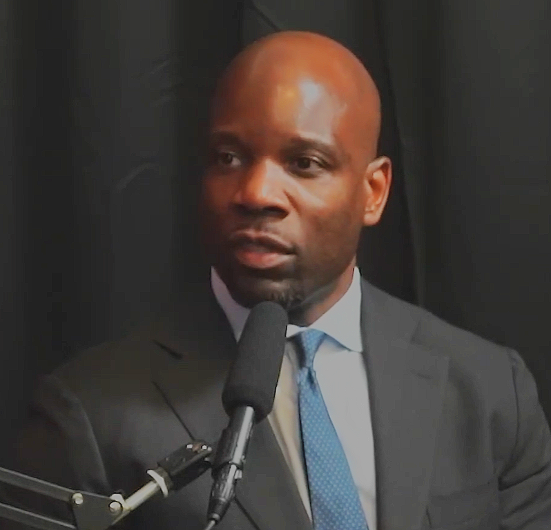 A man in a suit speaking into a microphone during an interview or speech.