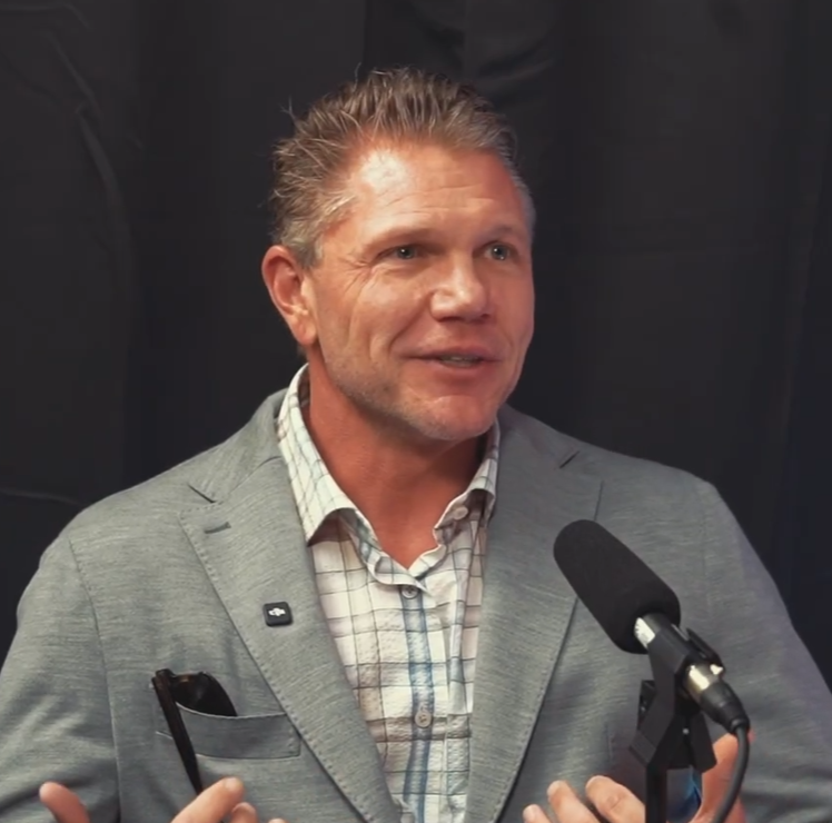 A man in a gray blazer and checkered shirt speaking into a microphone.