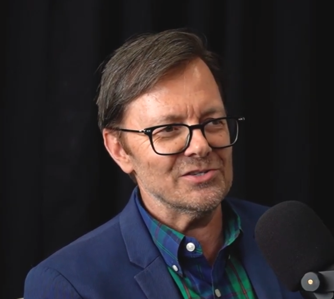 A man with glasses and brown hair, wearing a blue blazer and a green and blue checkered shirt, speaking into a microphone against a dark background.