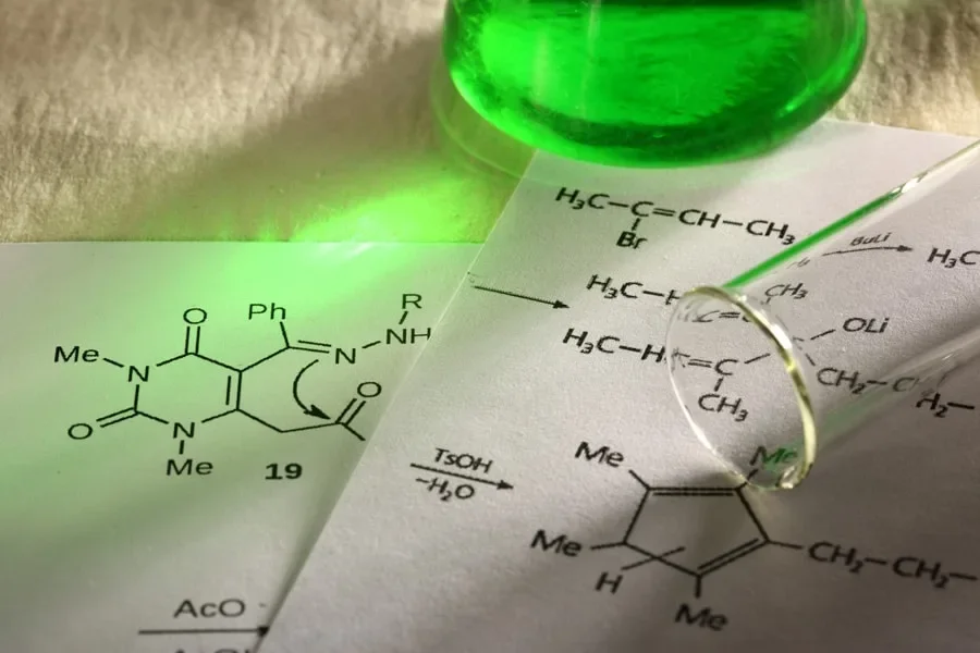 Laboratory paper with chemical structures, a green liquid in a glass container, and a magnifying glass highlighting part of the chemical diagram.