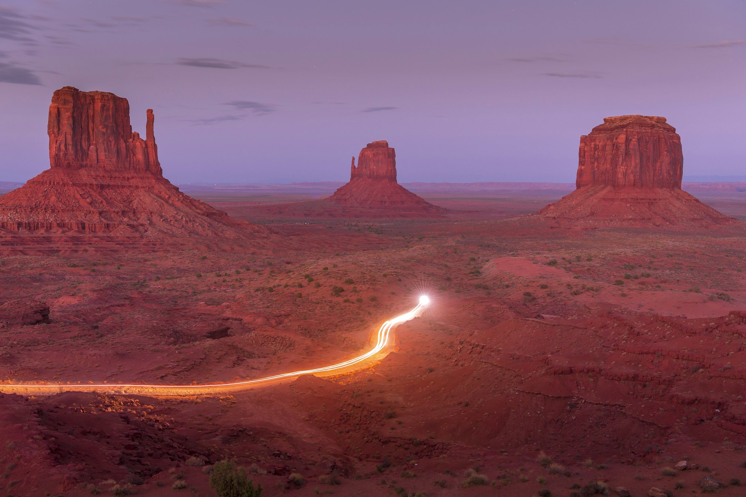 Sunset over three large red rock formations in Monument Valley, Arizona, with a trail of light from a moving vehicle in the foreground.