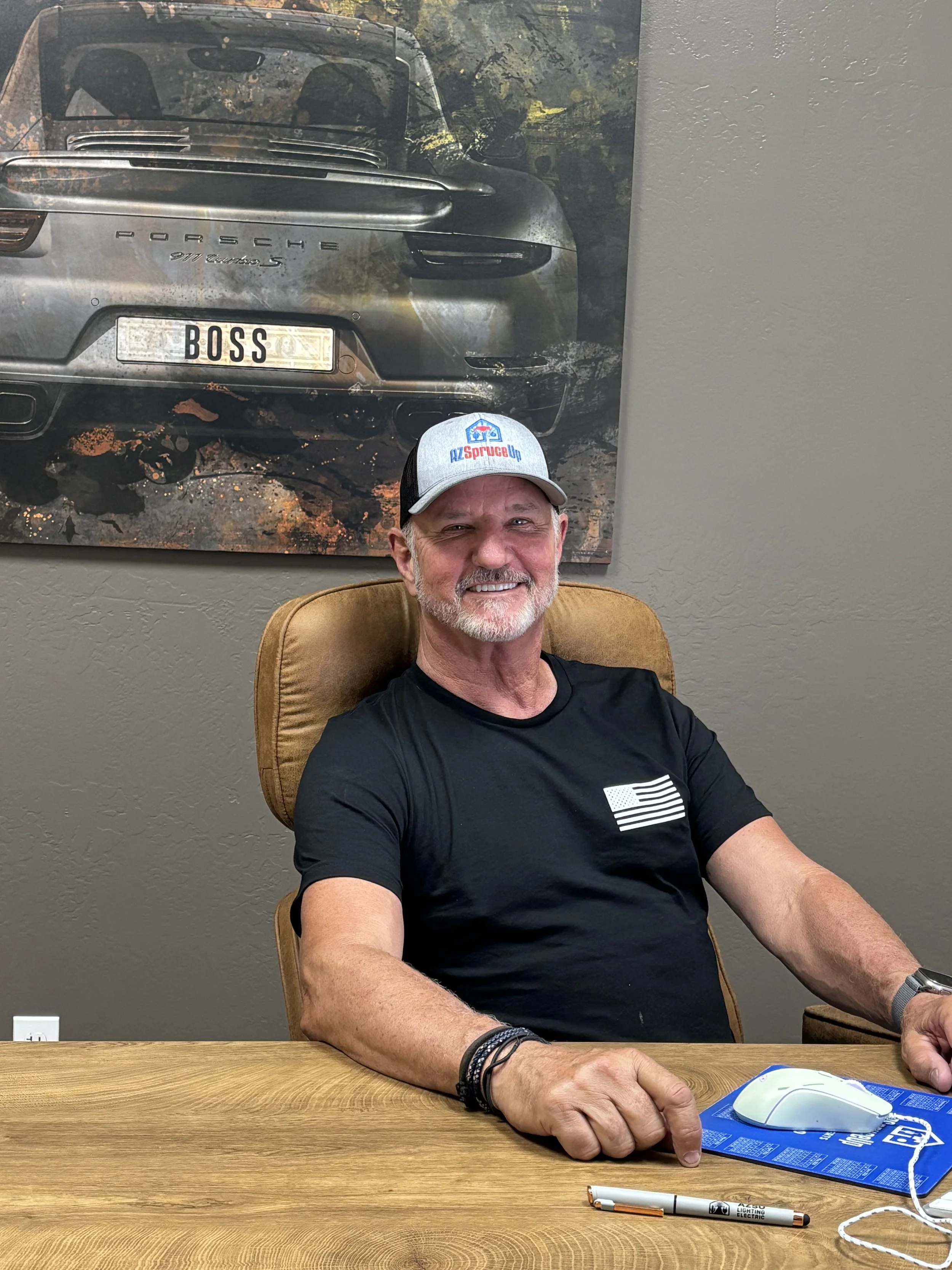 A smiling man with a beard and gray hair, wearing a gray cap with a logo, a black T-shirt with a white American flag, sitting at a wooden table in front of a gray wall, with a large photo of a silver Porsche car on the wall behind him.