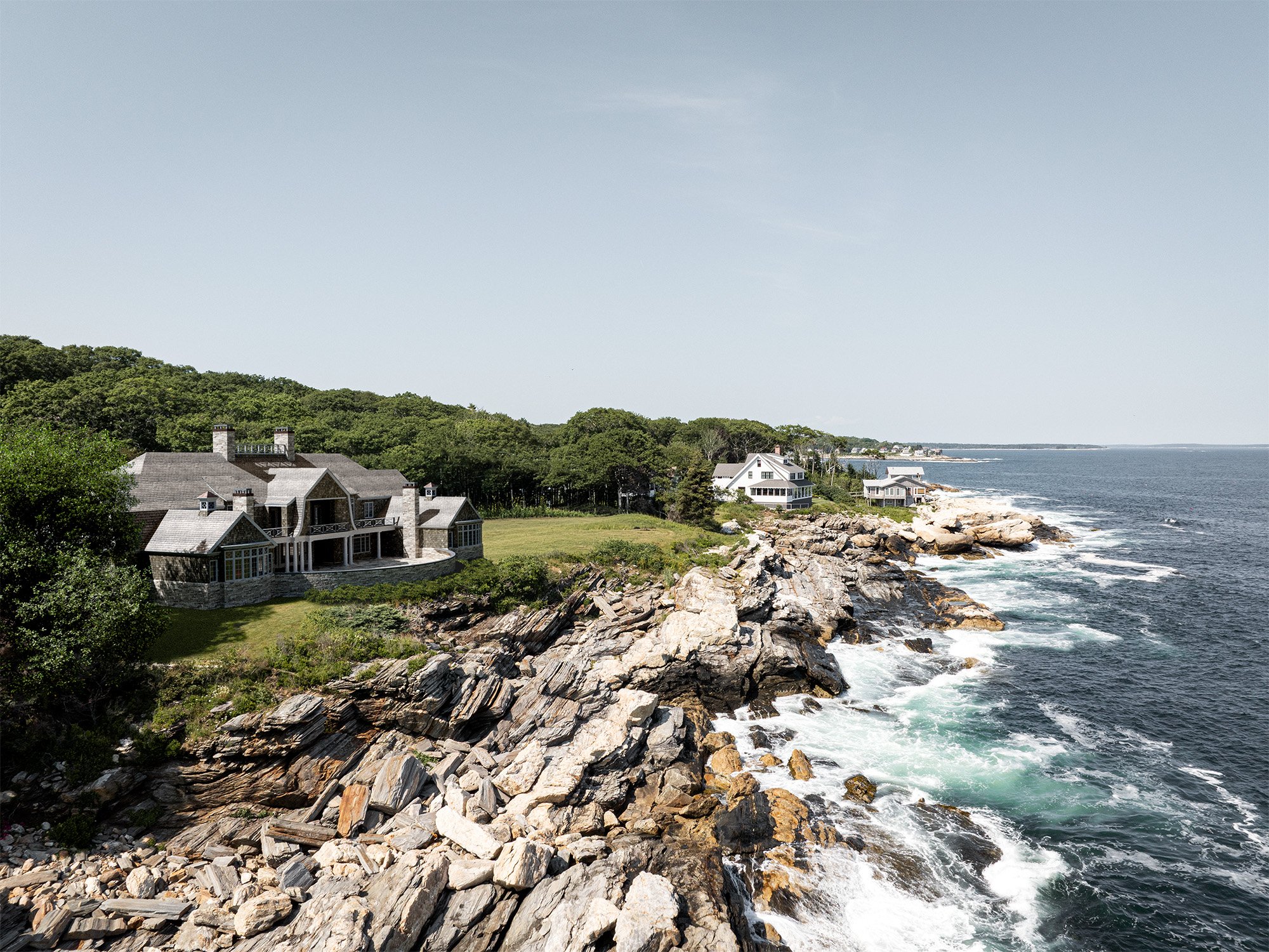 A coastal scene with large rocks along the shoreline, white waves crashing against them, and houses nestled among trees on a green hillside overlooking the ocean, with a clear blue sky in the background.