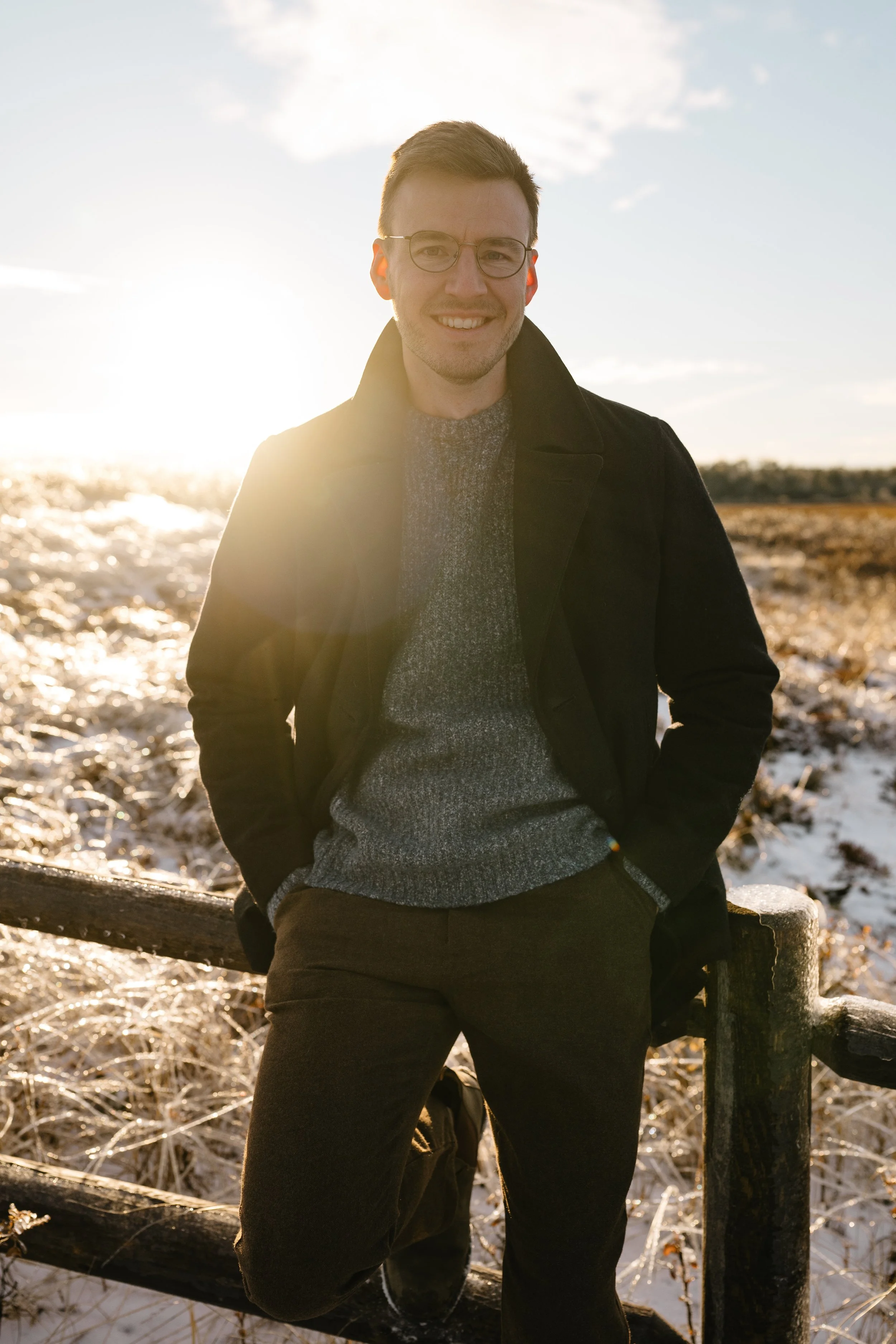 A young man smiling outdoors during sunset, dressed in a dark coat, glasses, and a gray sweater, standing next to a wooden fence with a winter landscape behind him.