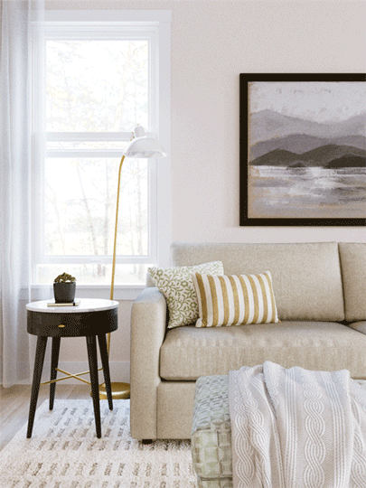 Living room with a beige sofa, striped pillows, a black side table with a plant, a floor lamp, a window with white curtains, a framed black-and-white landscape picture on the wall, and a cozy blanket on the sofa.