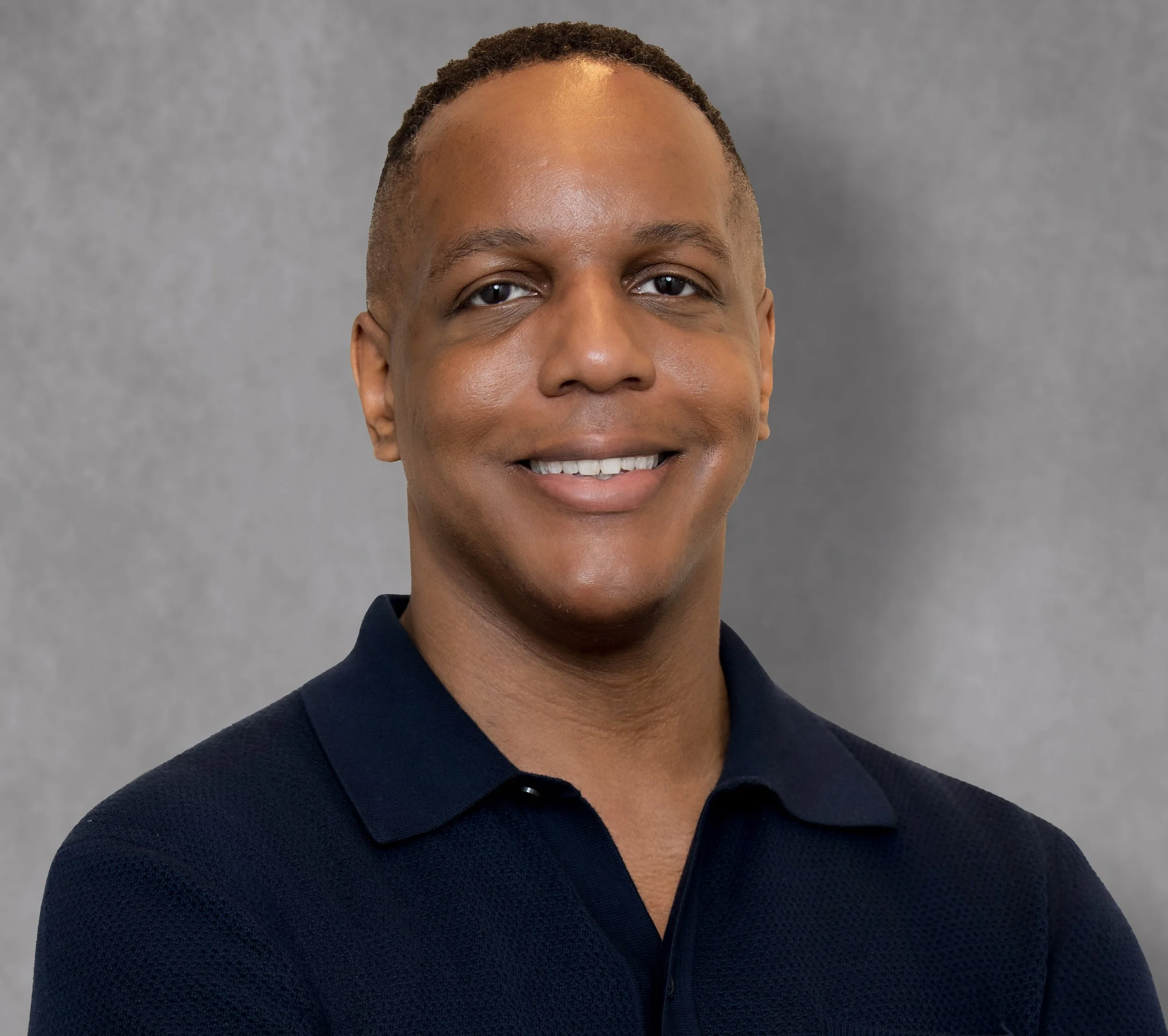 Close-up of a smiling person with short hair wearing a dark navy shirt against a gray background.
