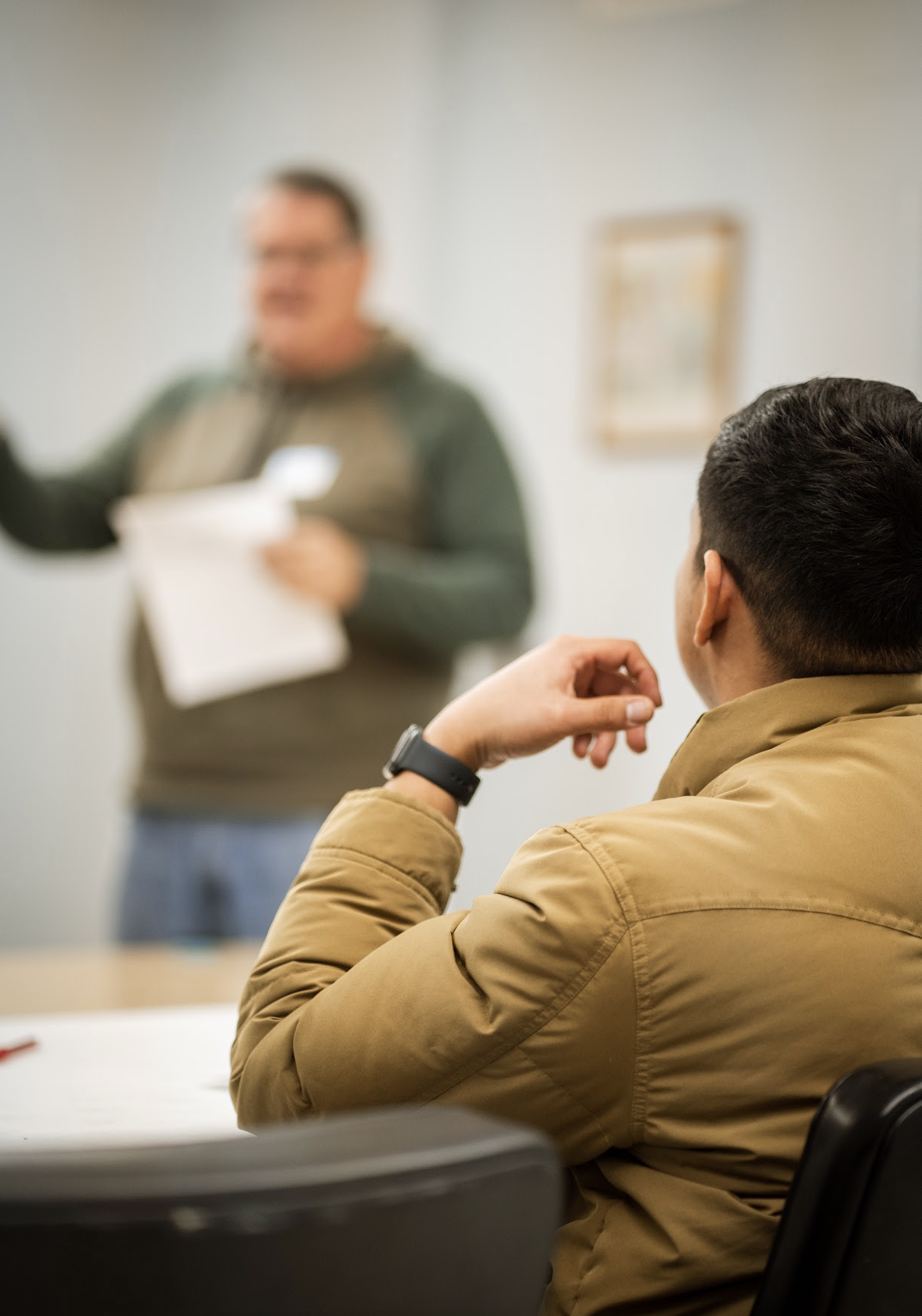 A person with short dark hair, wearing a tan jacket and a black watch, sitting at a table and listening attentively to a speaker in a classroom or meeting room. The speaker is a man with glasses, blurred out, holding a paper and gesturing.