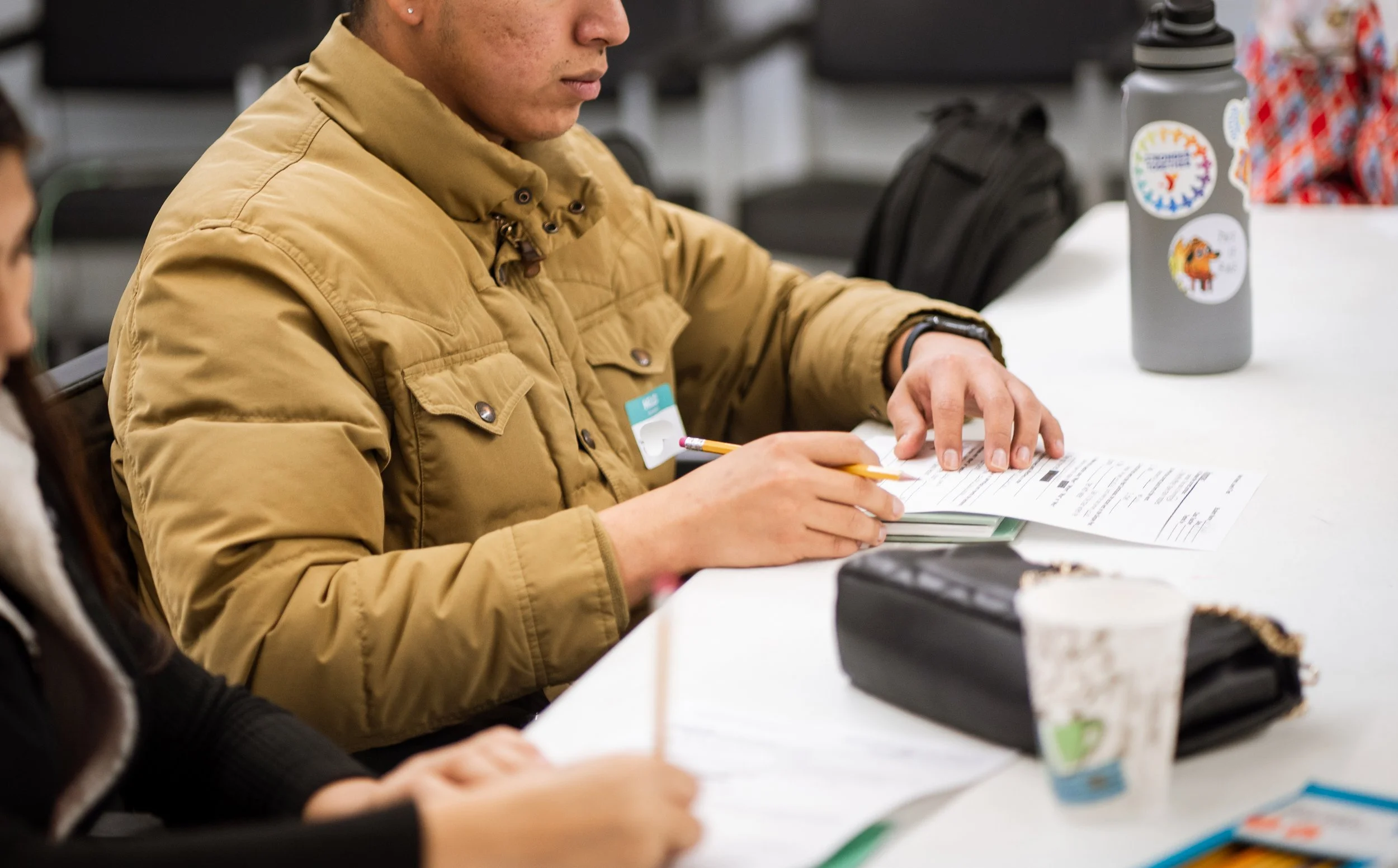 A person wearing a tan jacket, sitting at a table, writing on a document with a pencil. Several stickers are on a gray water bottle on the table.