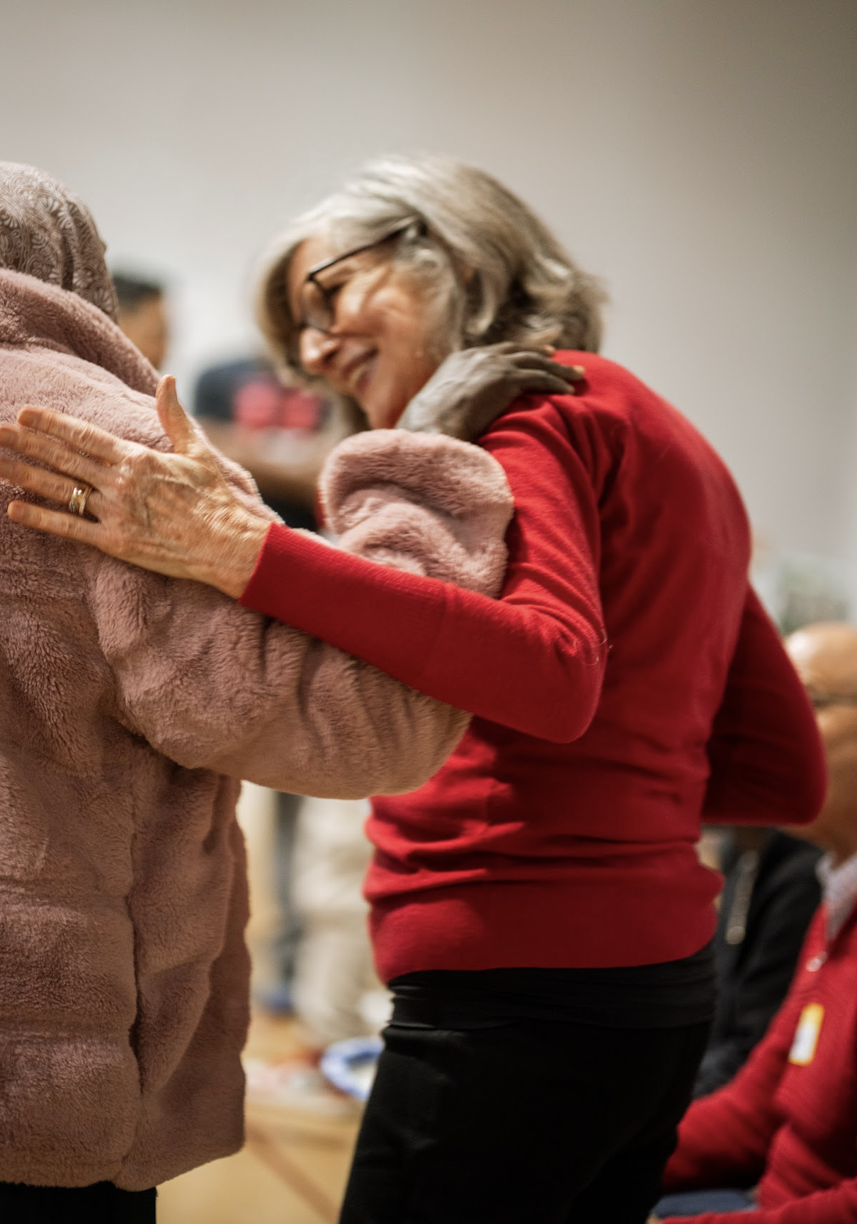 Two women hugging each other warmly, one wearing a pink fuzzy coat and the other wearing a red sweater and glasses, in an indoor setting.