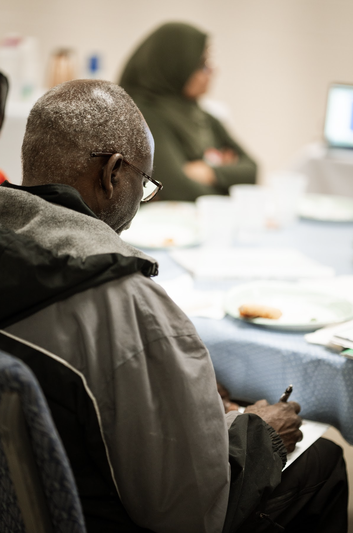 An elderly man with gray hair and glasses sitting at a table with others, using a smartphone.