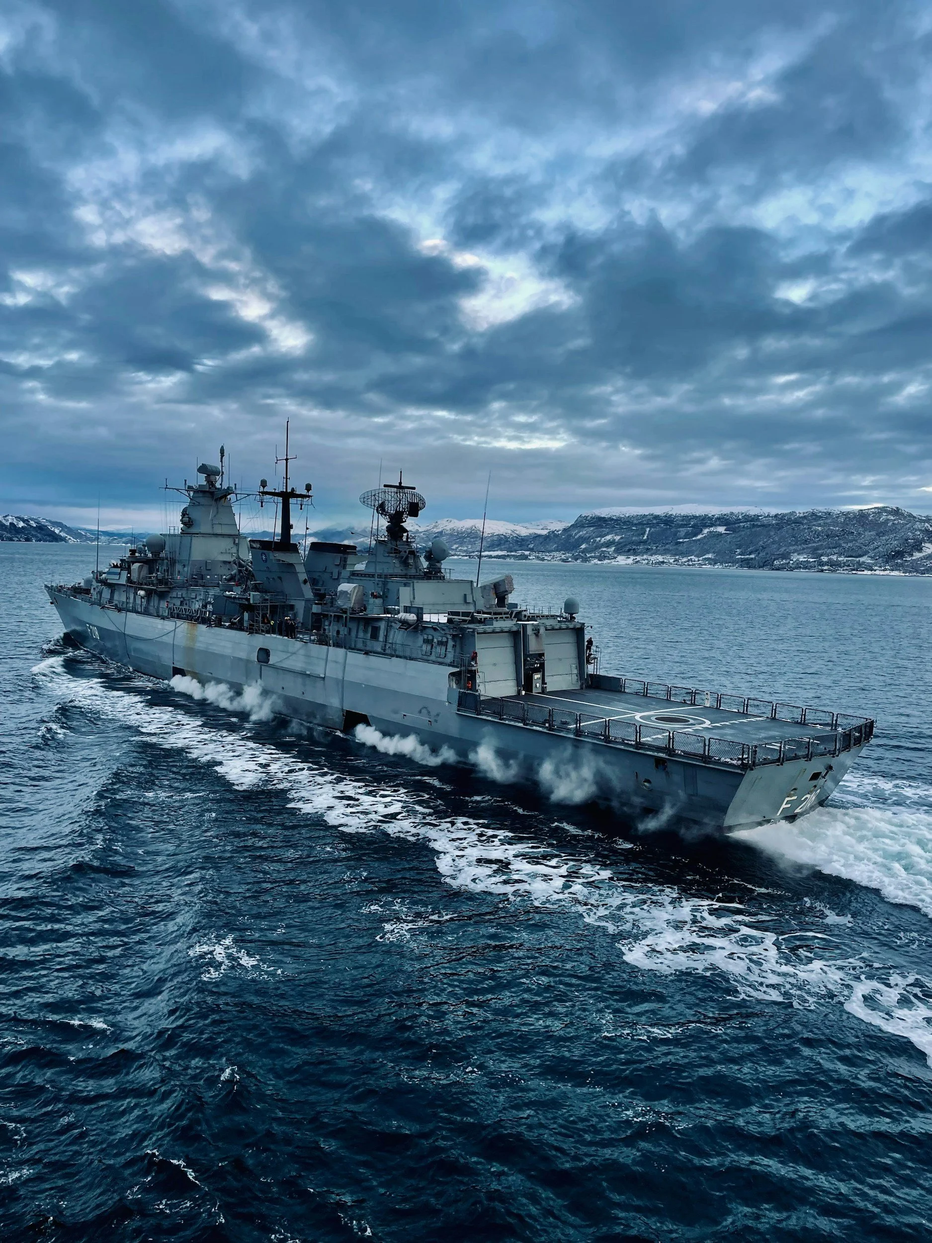 A military warship sailing through the sea with snow-covered mountains in the background under a cloudy sky.