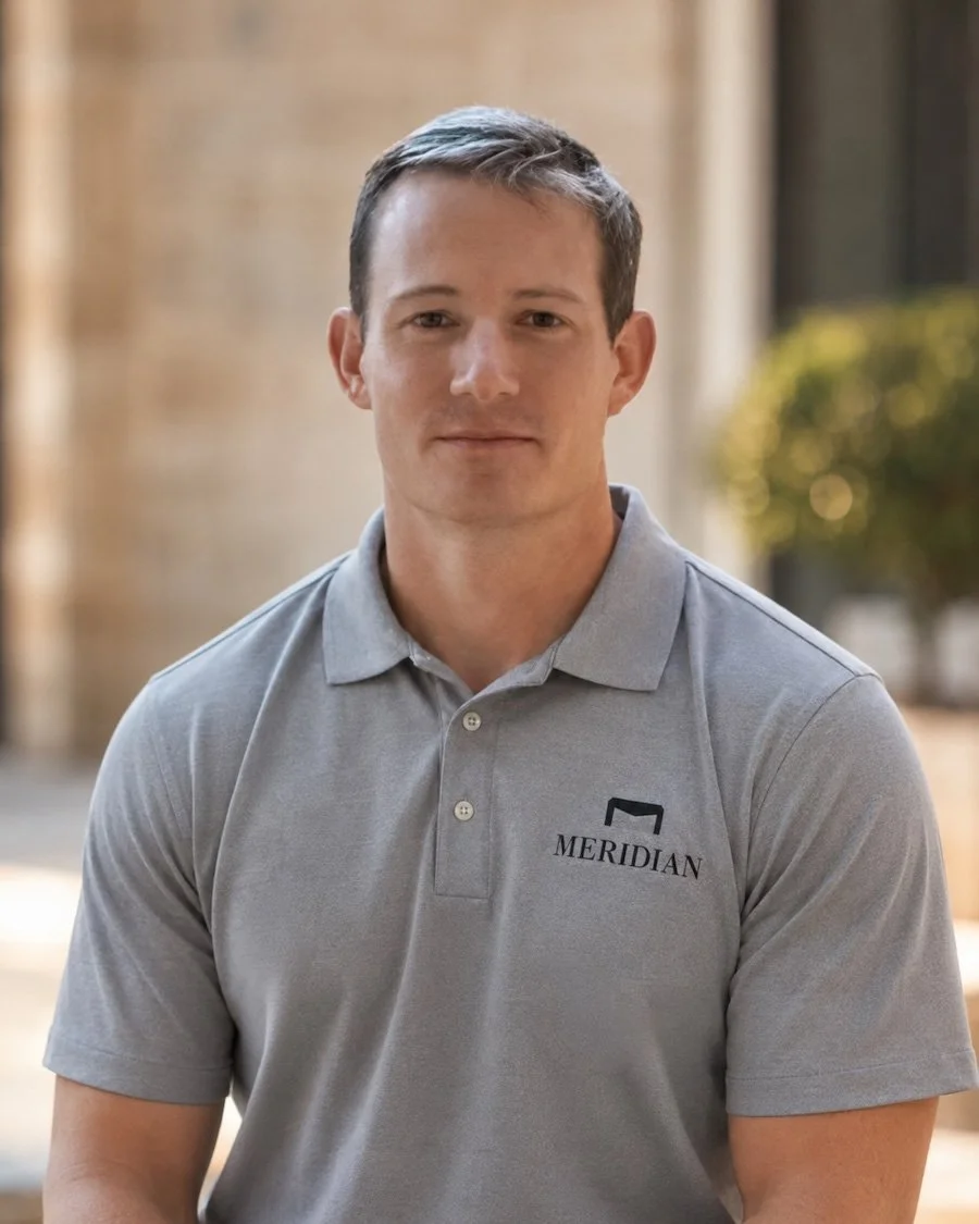 A young man with short brown hair wearing a gray polo shirt with the 'Meridian' logo, standing outdoors with a blurred background of trees and buildings.