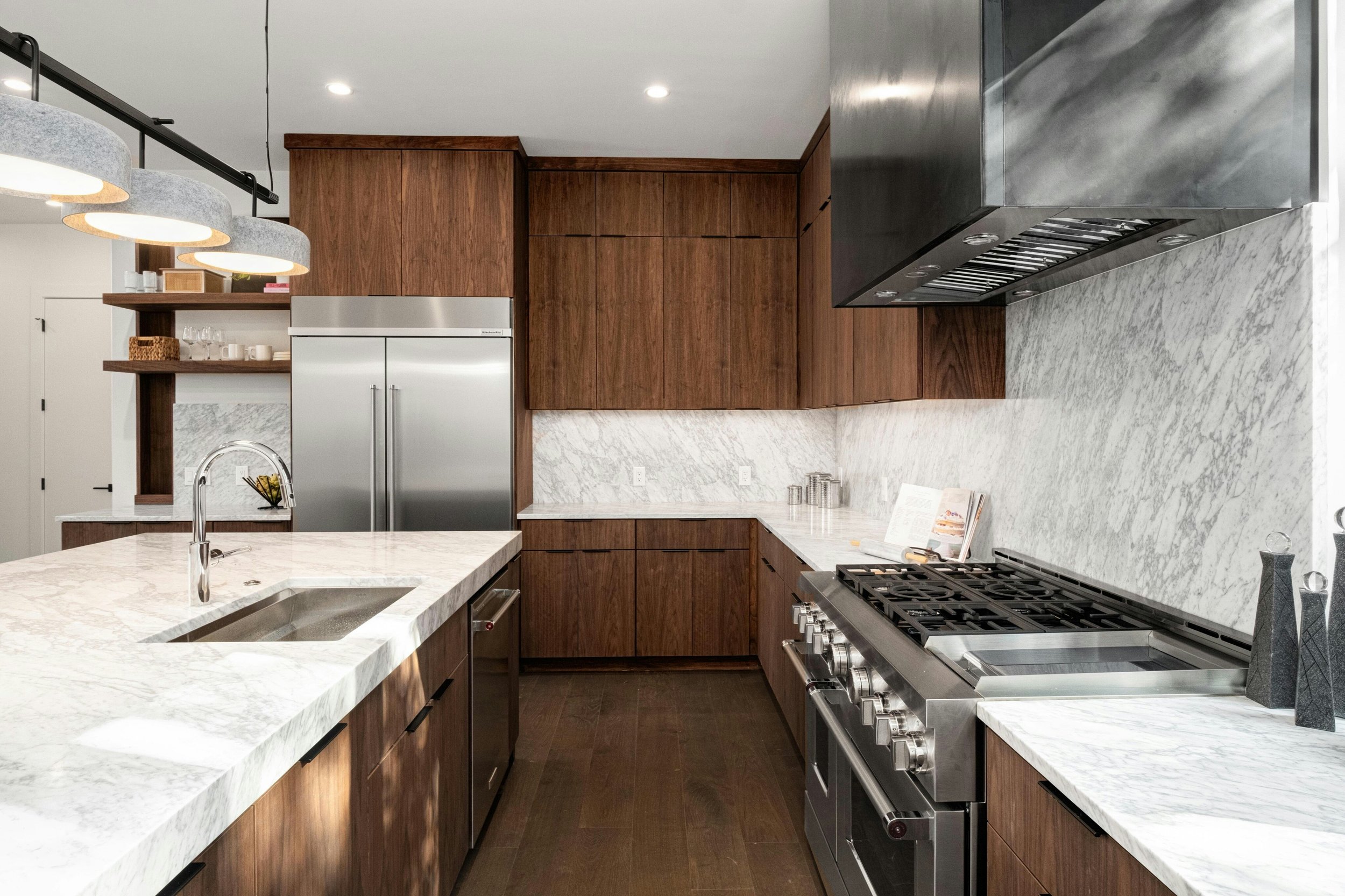Modern kitchen with white marble countertops, wooden cabinets, stainless steel appliances including a refrigerator and oven, and decorative pendant lights.
