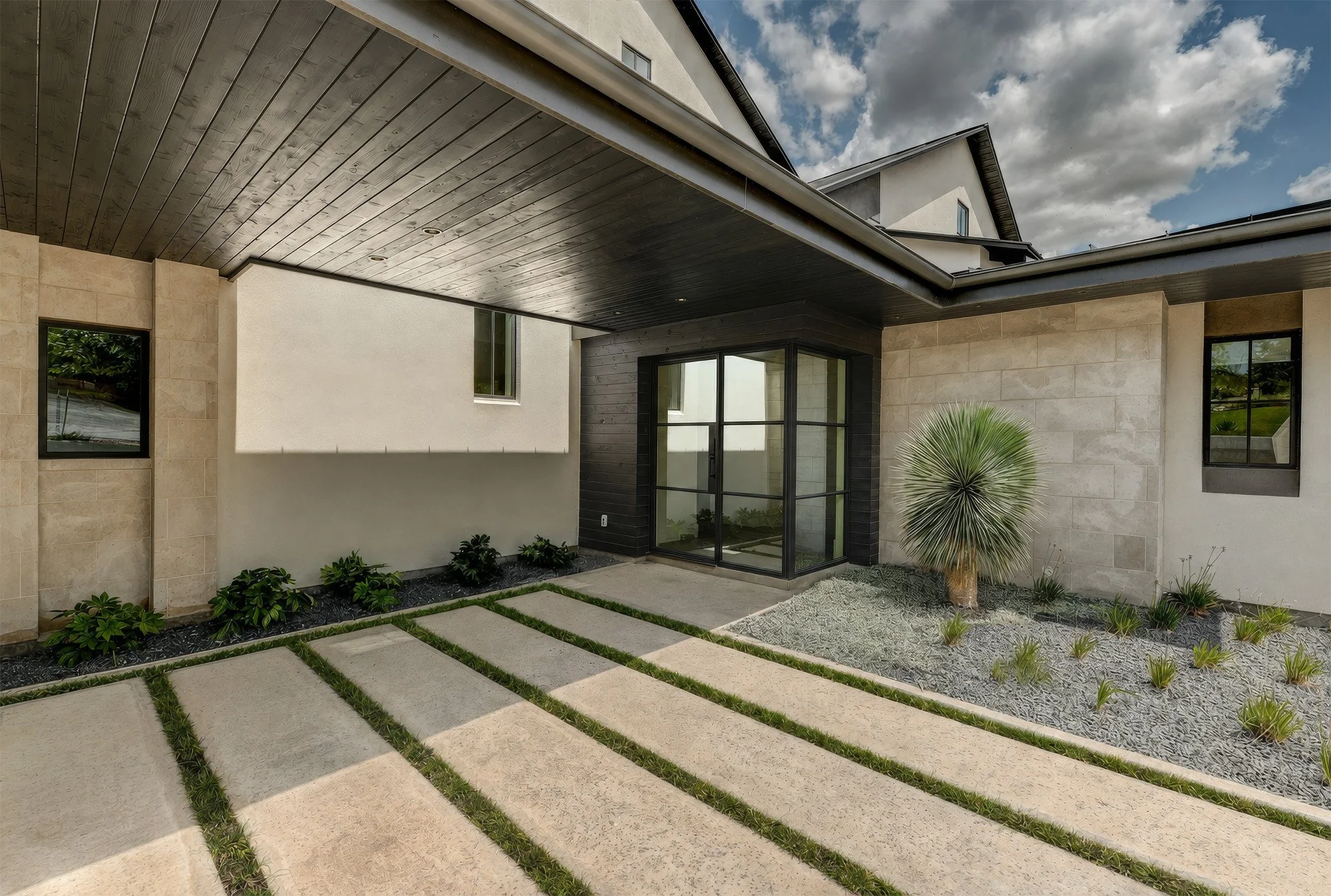 Exterior view of a modern house with a front yard featuring concrete stepping stones and landscaped plants. The house has beige walls, large windows, and black trim. The sky is partly cloudy.