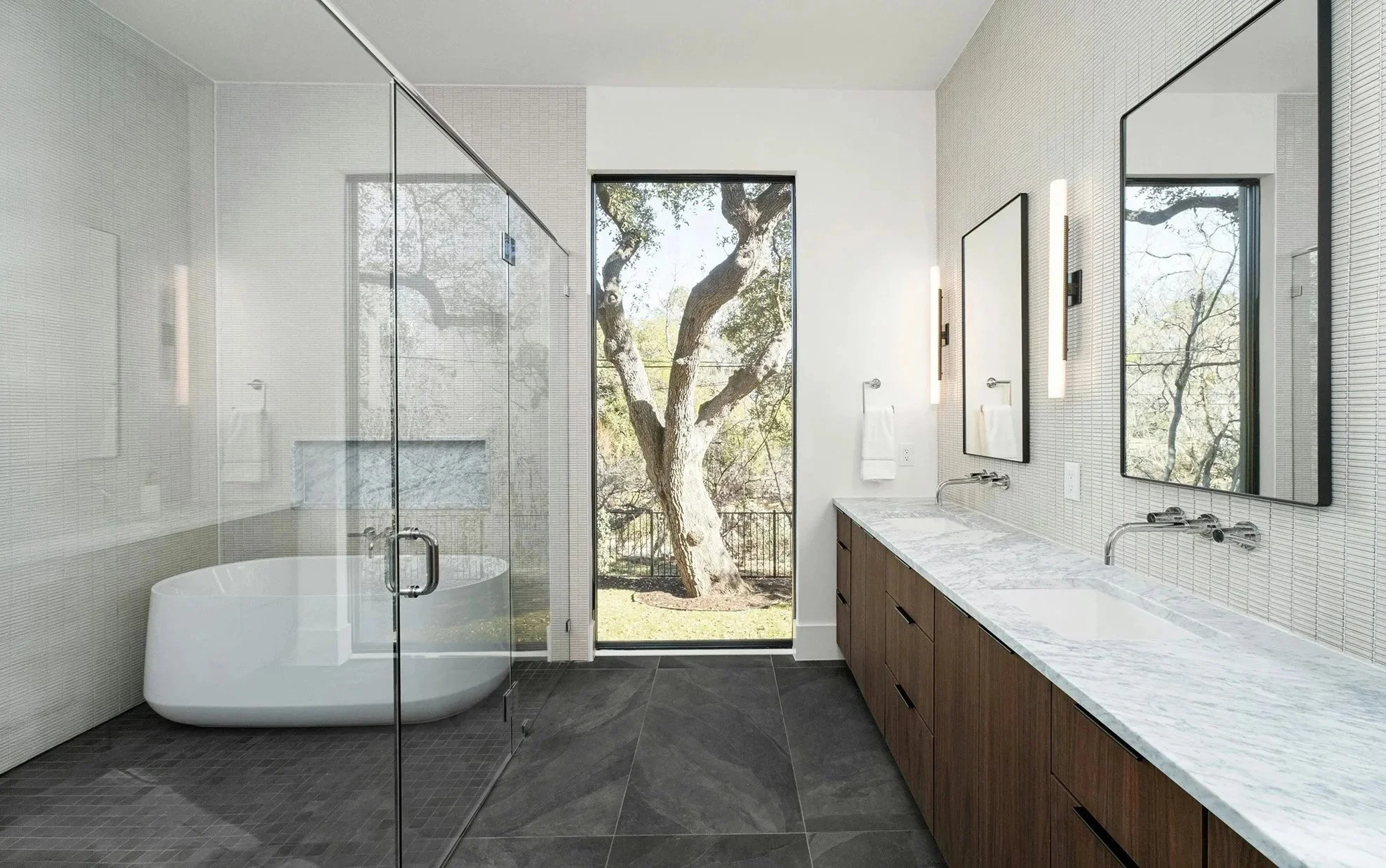 Modern bathroom with a walk-in shower and a large window overlooking a tree outside. There is a white bathtub, a marble countertop with a sink, and two mirrors on the wall.