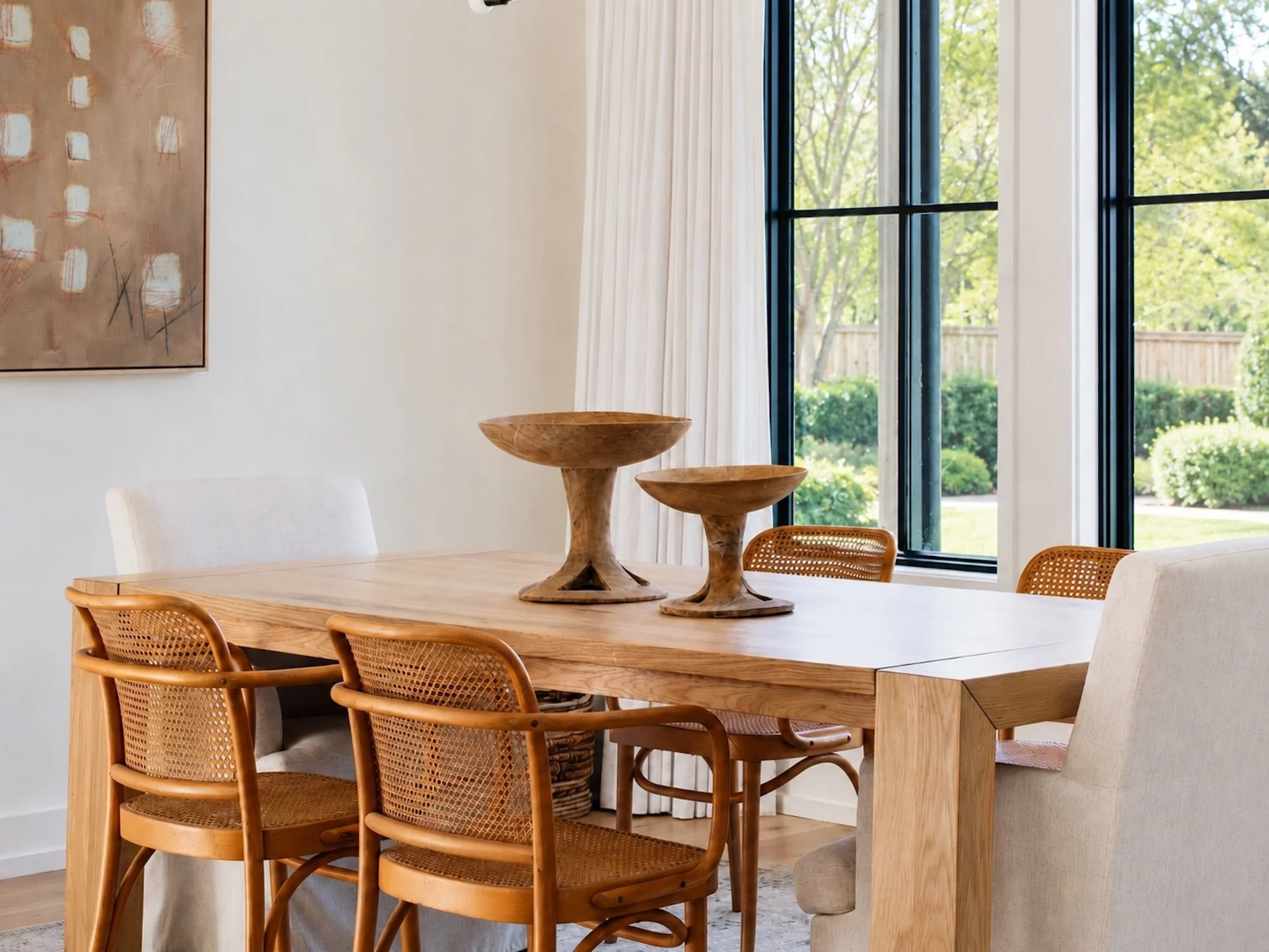 A dining room with a wooden table, rattan chairs, and modern decor, with large windows revealing a green outdoor area.