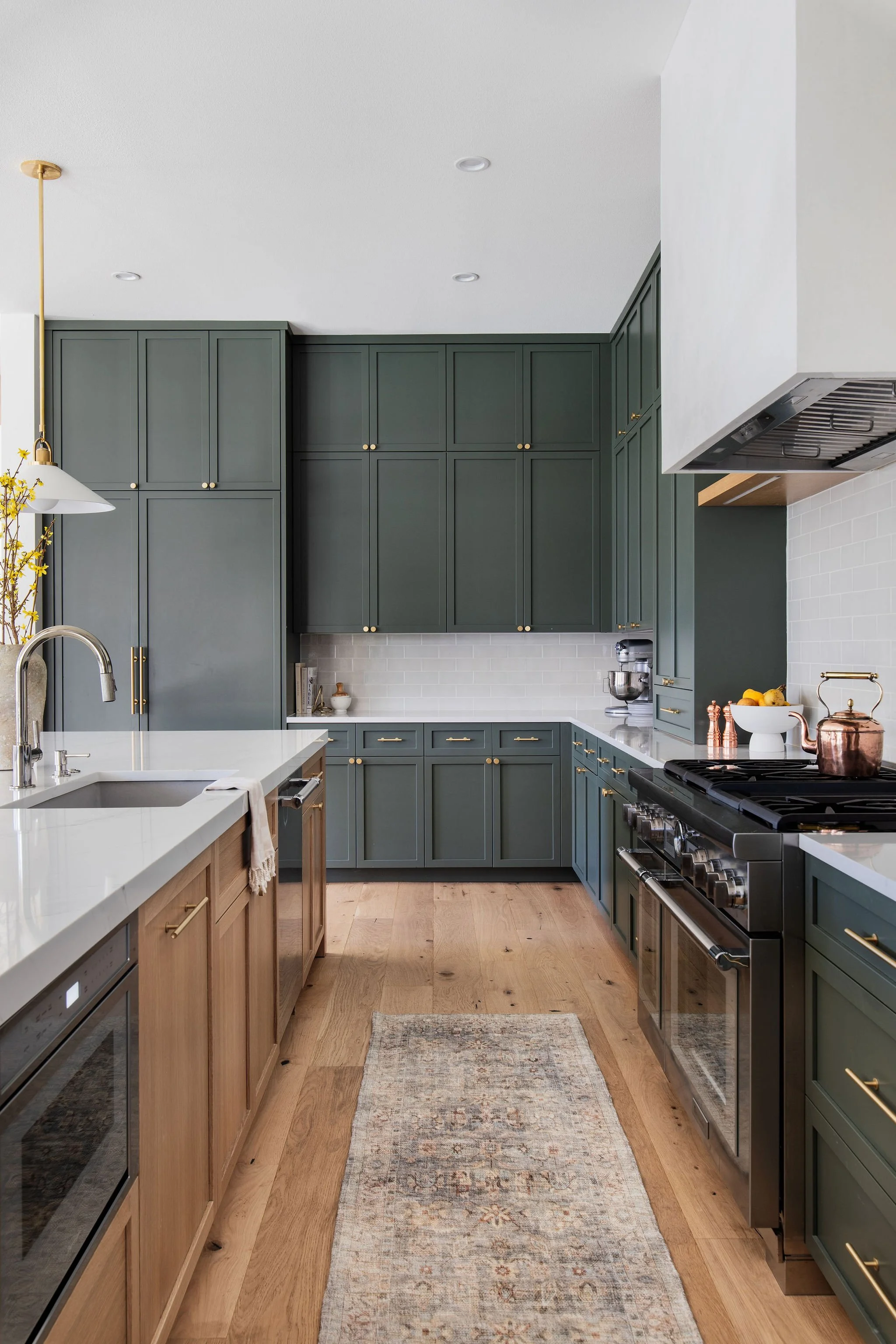 A modern kitchen with green cabinets, white subway tile backsplash, a kitchen island with a light wood base and white countertop, and a stainless steel stove. The floor is light wood, and there is a small rug in the center.