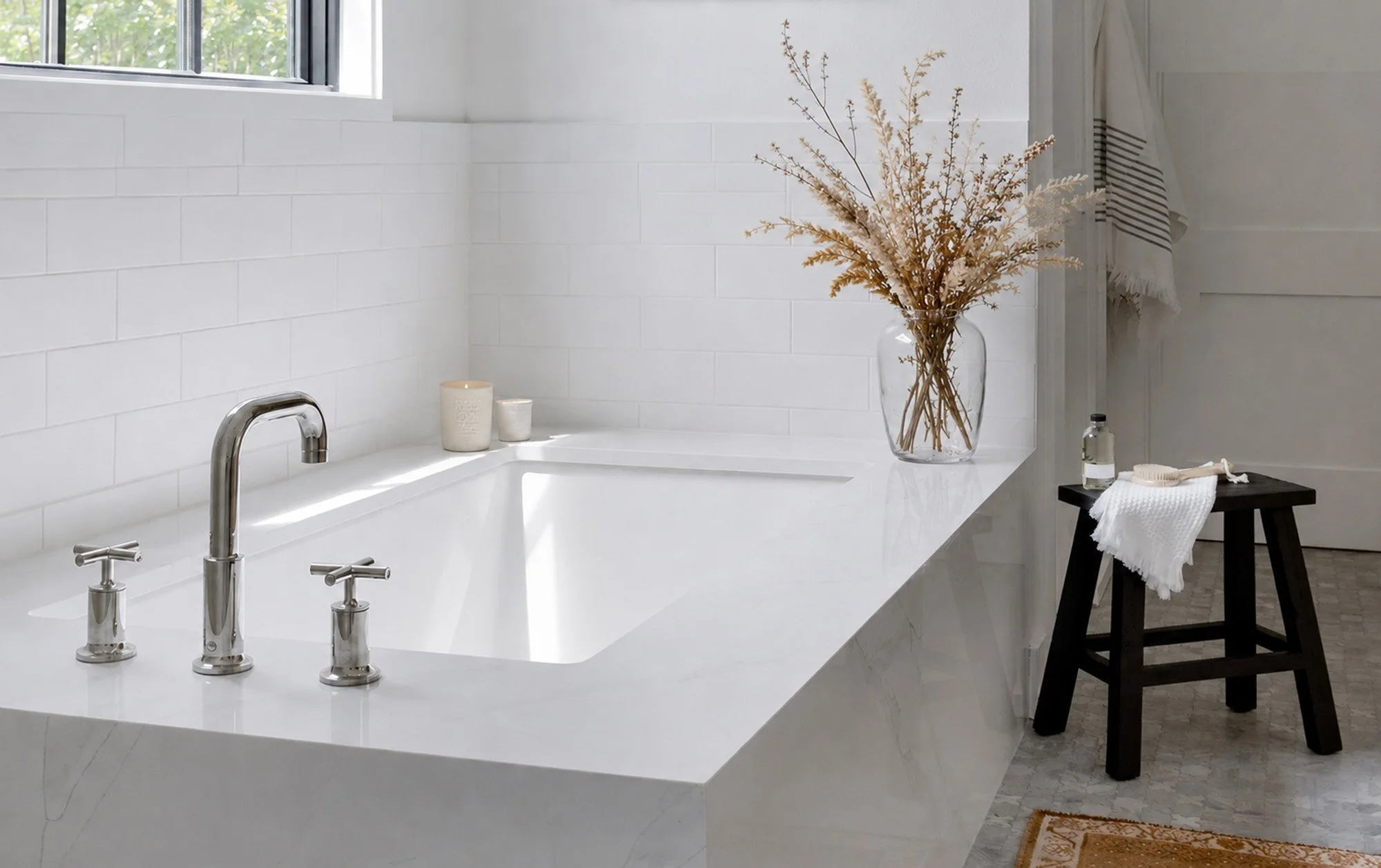 A bathroom with a white bathtub, a window above it, and a clear glass vase with dried flowers on the side. There are candles and a towel on a small black stool nearby.