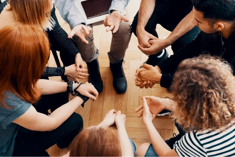 Group of people sitting in a circle, holding hands, engaging in a support group.