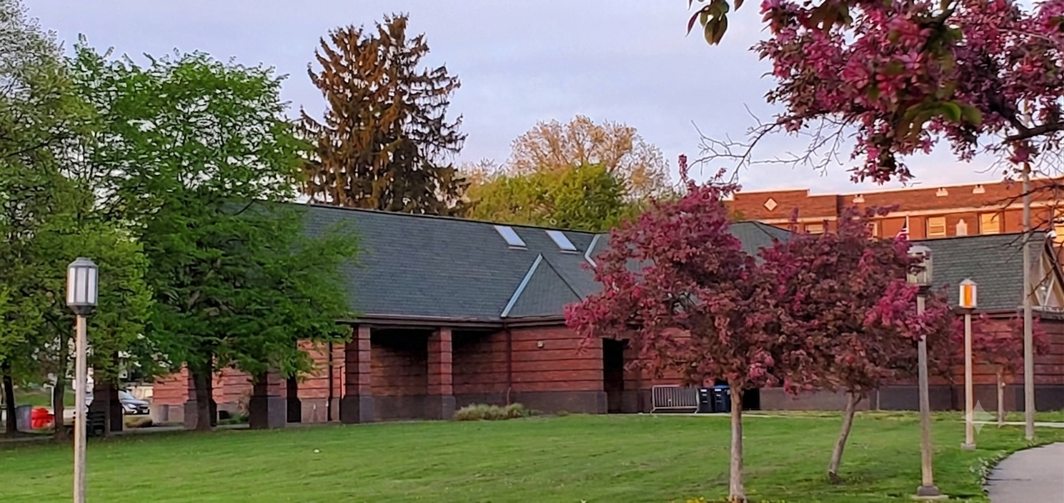 A park with green grass, pink and green trees, and a building with a gray roof in the background. There are also several lampposts along a sidewalk.