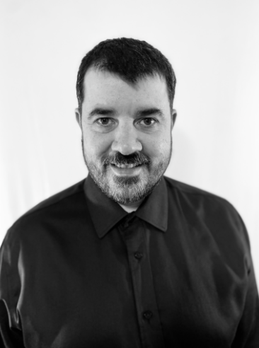 Black-and-white head-and-shoulders portrait of a smiling bearded man wearing a black button-up shirt.